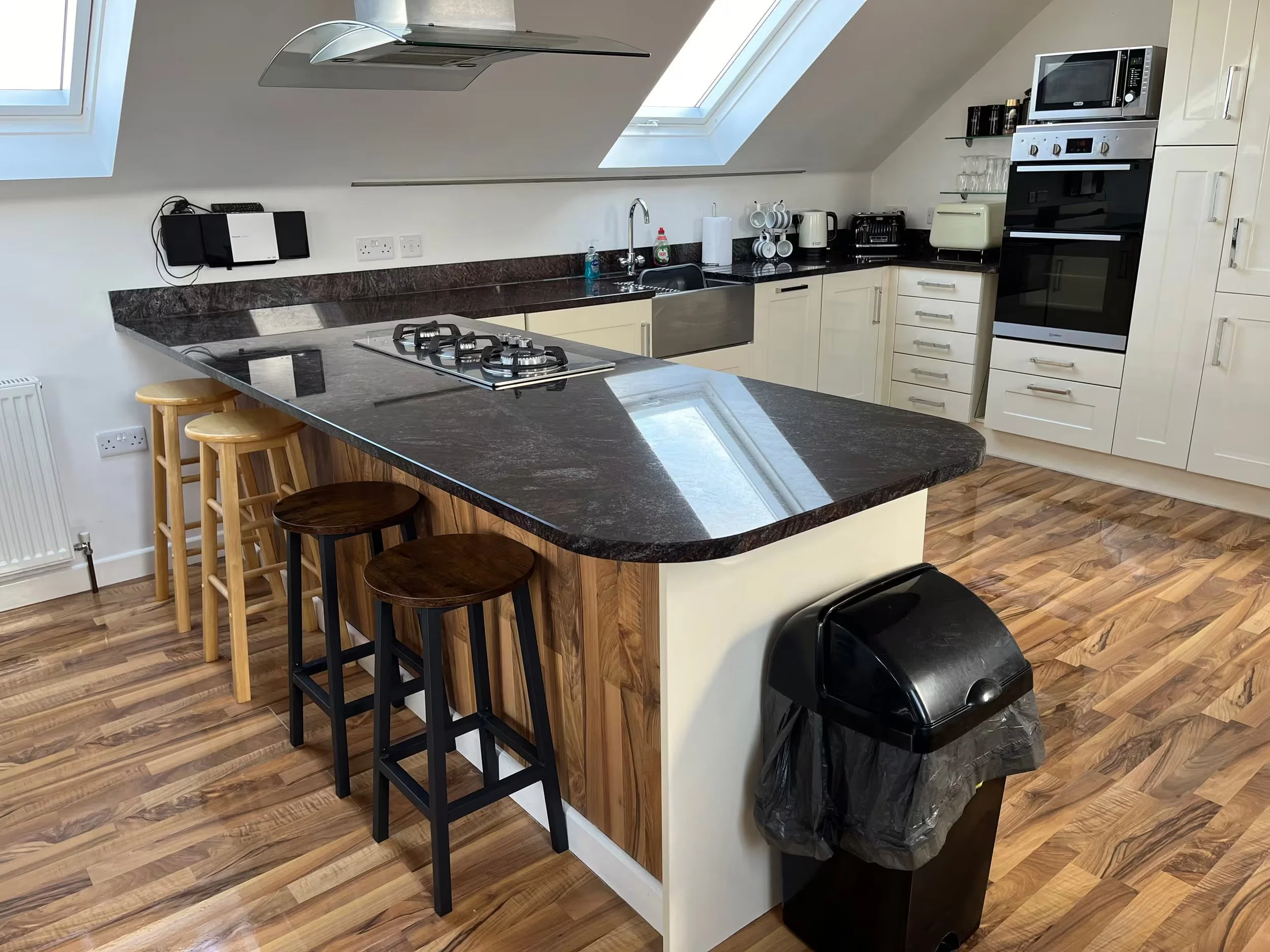 Modern kitchen at Sunnyridge with skylight windows, dark granite countertop, cream-colored cabinets, and wood flooring. Contains stove, microwave, oven, toaster, electric kettle, and hanging cups, with a small breakfast bar and bar stools.