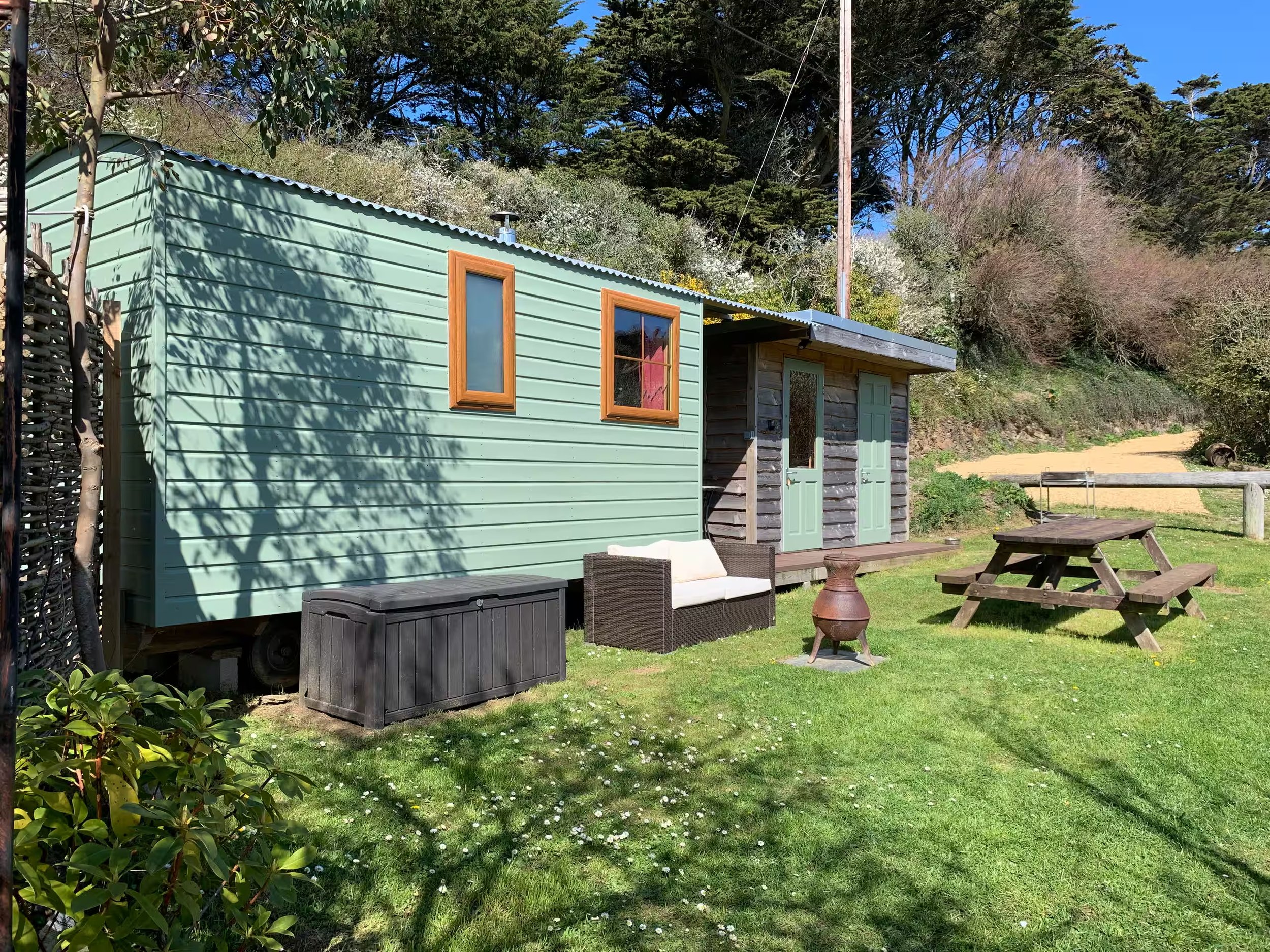Foxes Cove shepherds hut with two windows and green doors, surrounded by a grassy garden with outdoor furniture, against a backdrop of trees and a clear blue sky.