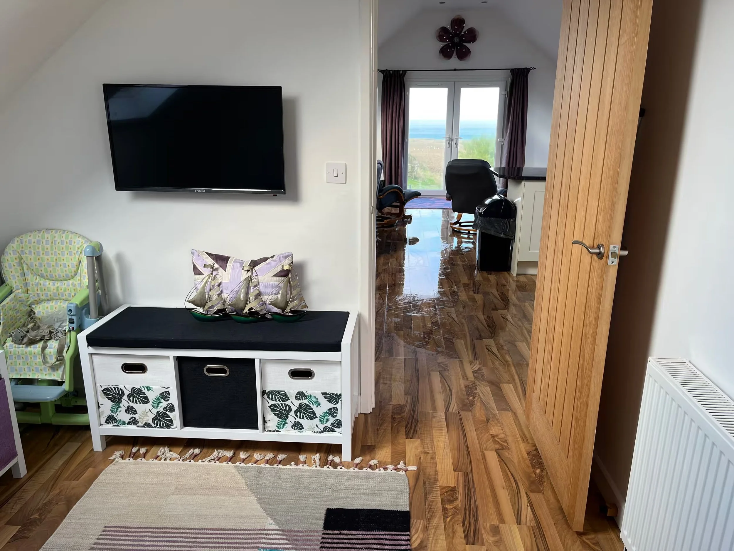 Interior of snug room at Sunnyridge with a flat-screen television mounted on a white wall, a child's high chair, a white storage unit, a rug, and a view through a doorway of a room with a view of the ocean and two chairs facing the window.