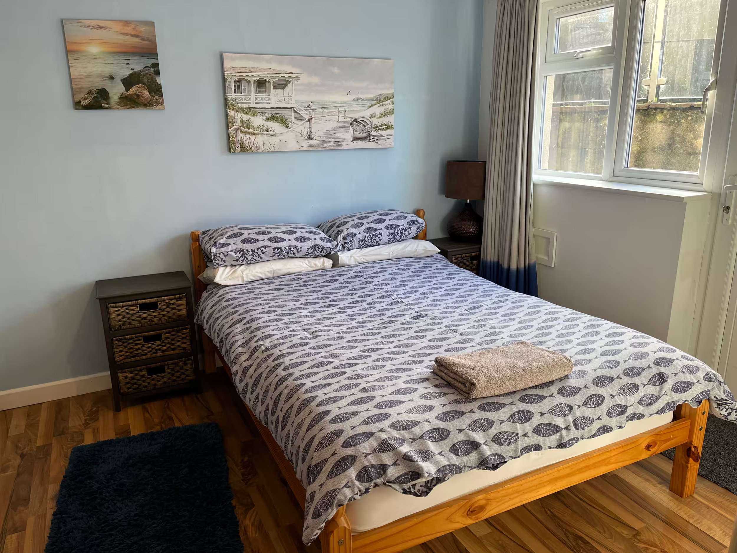 The double bedroom with nightstand and wicker basket drawer and a brown lamp beside the bed. The wall is decorated with two beach-themed paintings, A window with white frames and beige curtains lets in natural light, revealing the outdoor greenery.