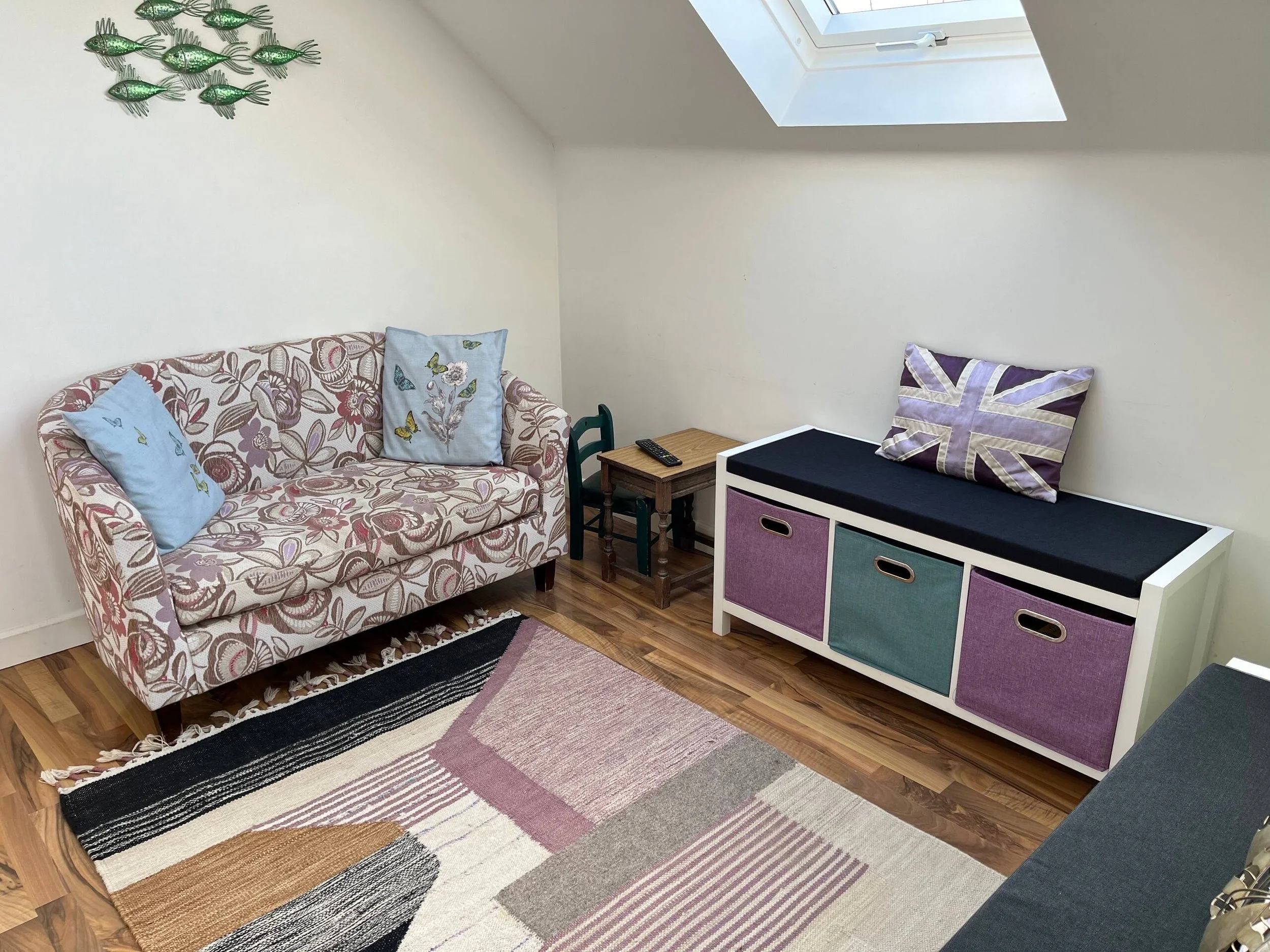 A cozy snug area with a sofa at Sunnyride.  There's a small wooden side table with a TV remote, and a multi-colored storage. The room has a skylight window, a colourful striped rug on hardwood floors, and wall decorations of green fish-shaped art.