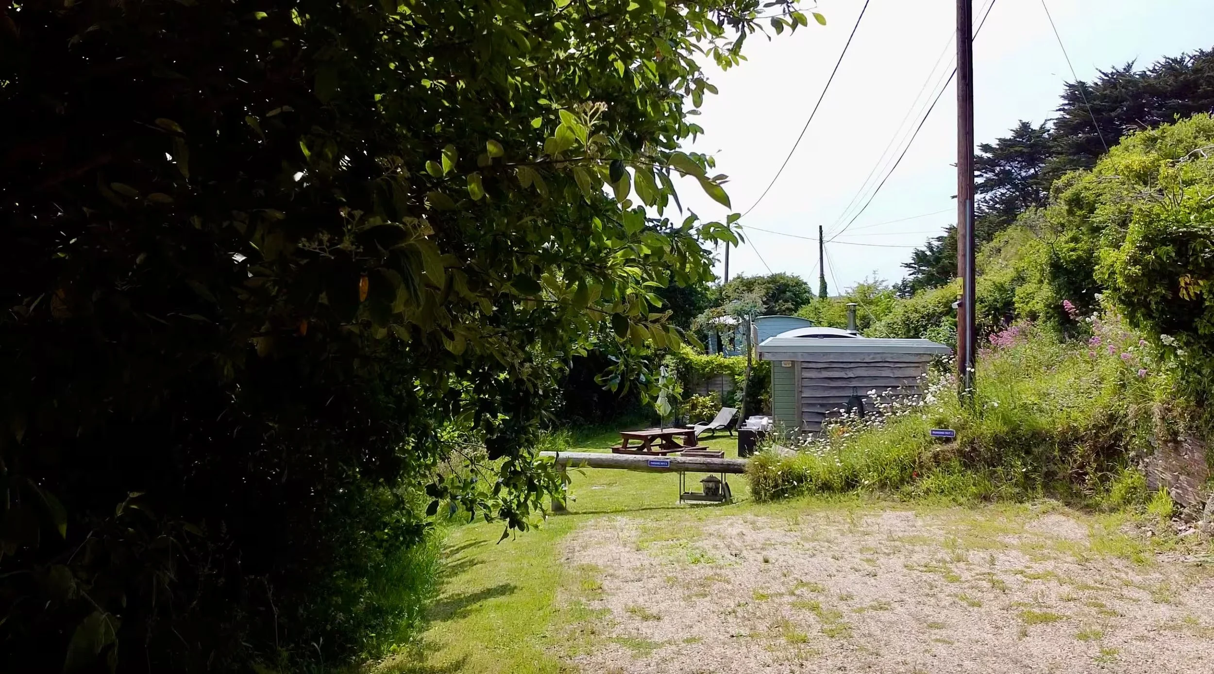 Parking at Porthcothan Retreats with garden with lush green trees and shrubs, a sitting area with a picnic table and chairs, a small shed, and flowering plants along a pathway, under a bright sky with power lines.
