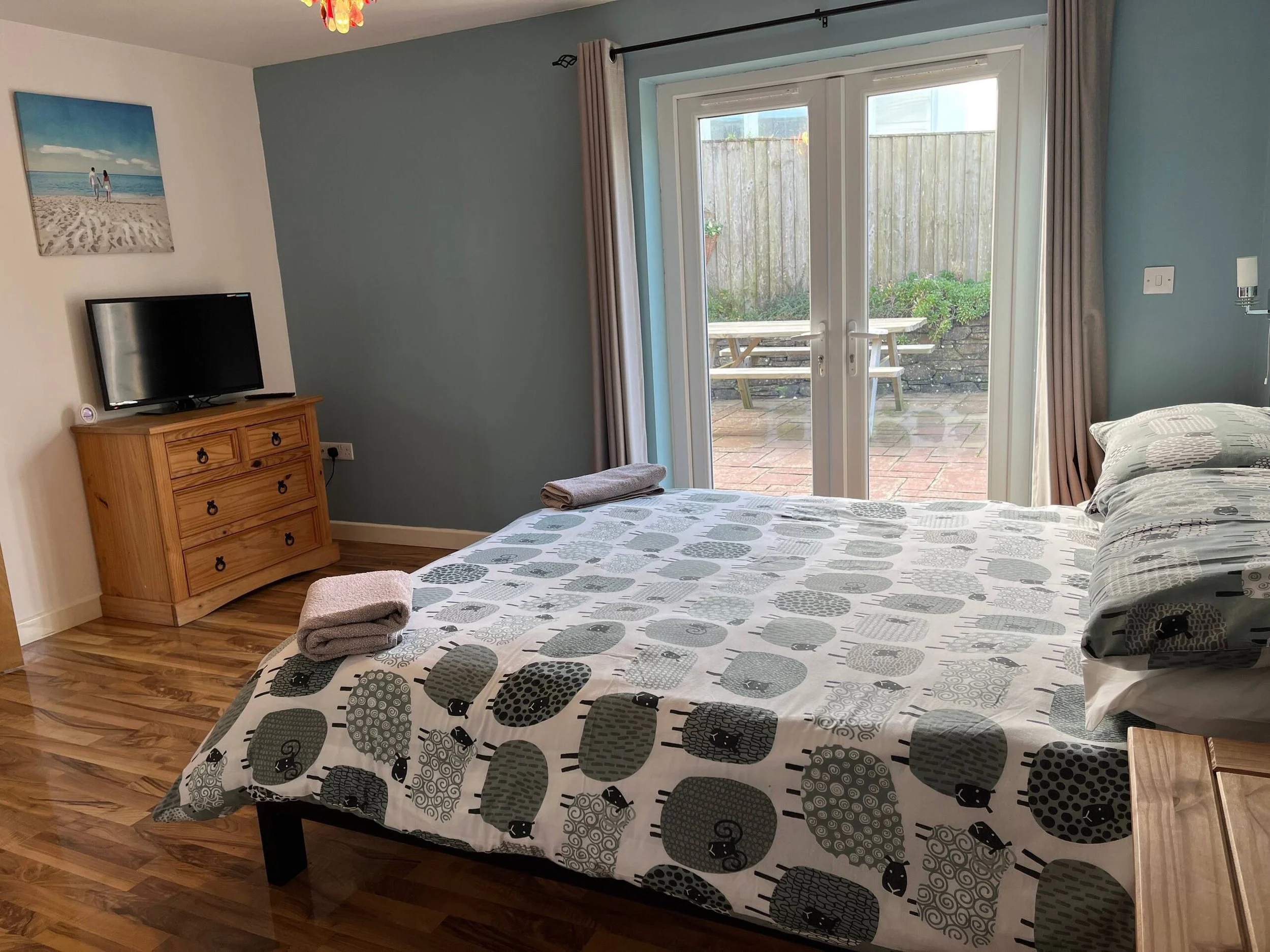 A bedroom with a bed covered in a black and gray pillowcase, a small wooden dresser with a flat-screen TV on top, a wall painting of two people holding hands on a beach, and glass doors leading to the outside patio.