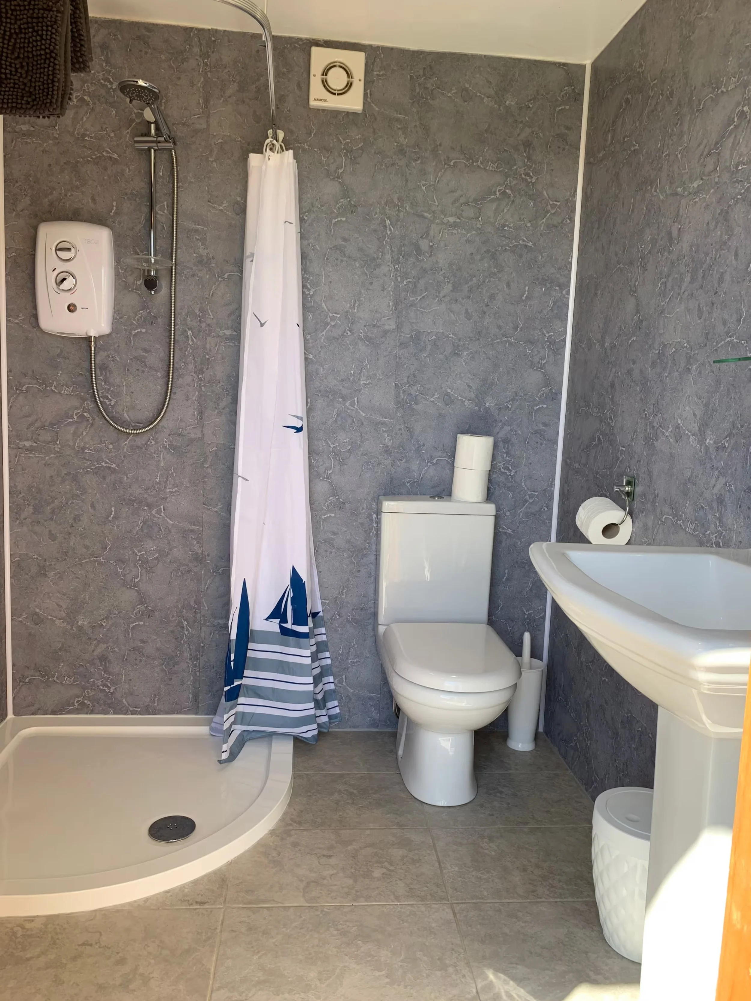 Foxes Cove bathroom with grey tiled walls and floor, a white shower with a curtain, a white toilet, a wall-mounted sink, a soap or lotion dispenser, a toilet paper roll, a small trash can, and a small ventilation fan on the wall.