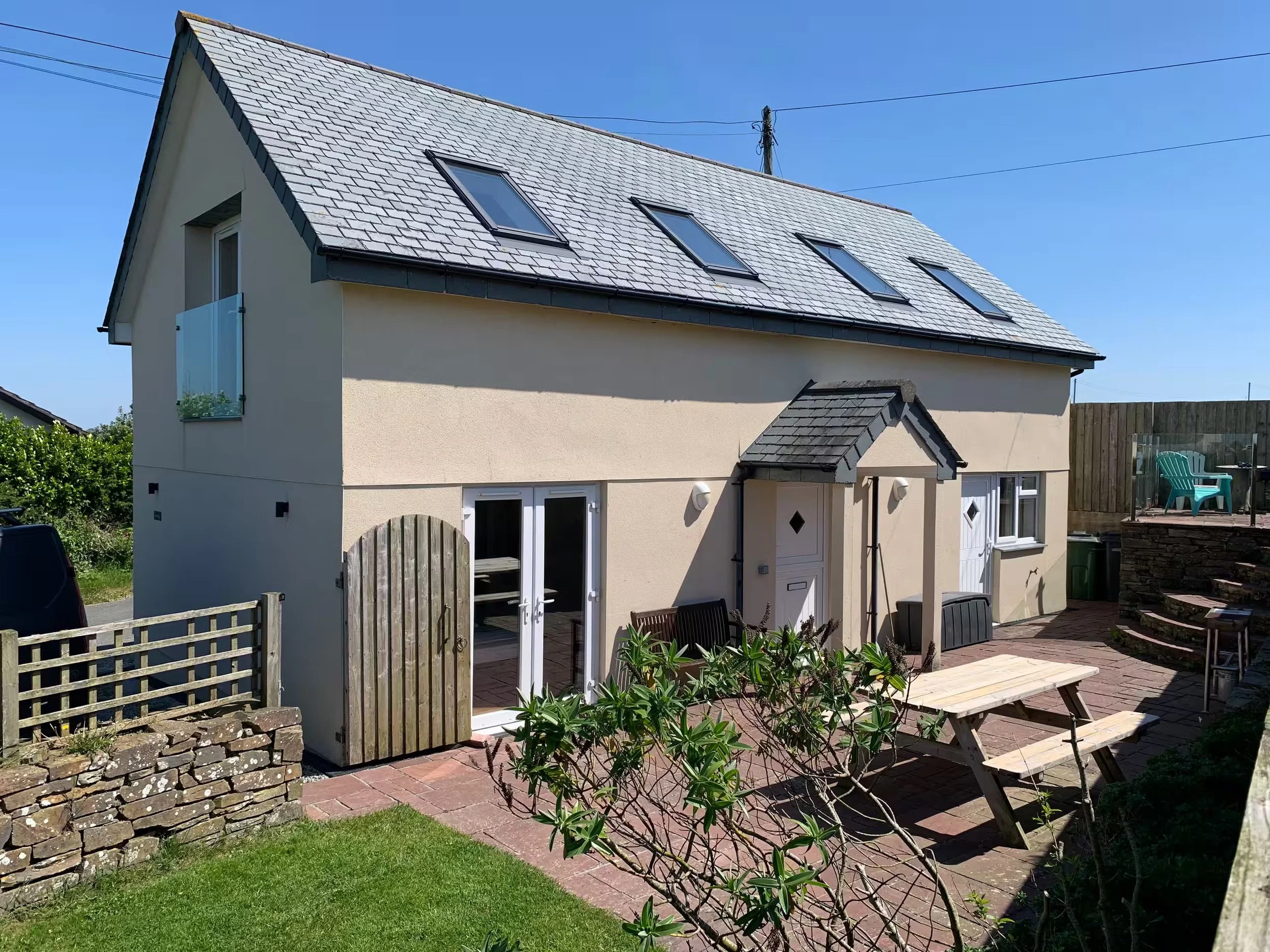 Sunnyridge patio area with a wooden picnic table, lush green plants, a small gate, brick flooring. The house with a sloped roof fitted with four skylights and a window with a flower box.