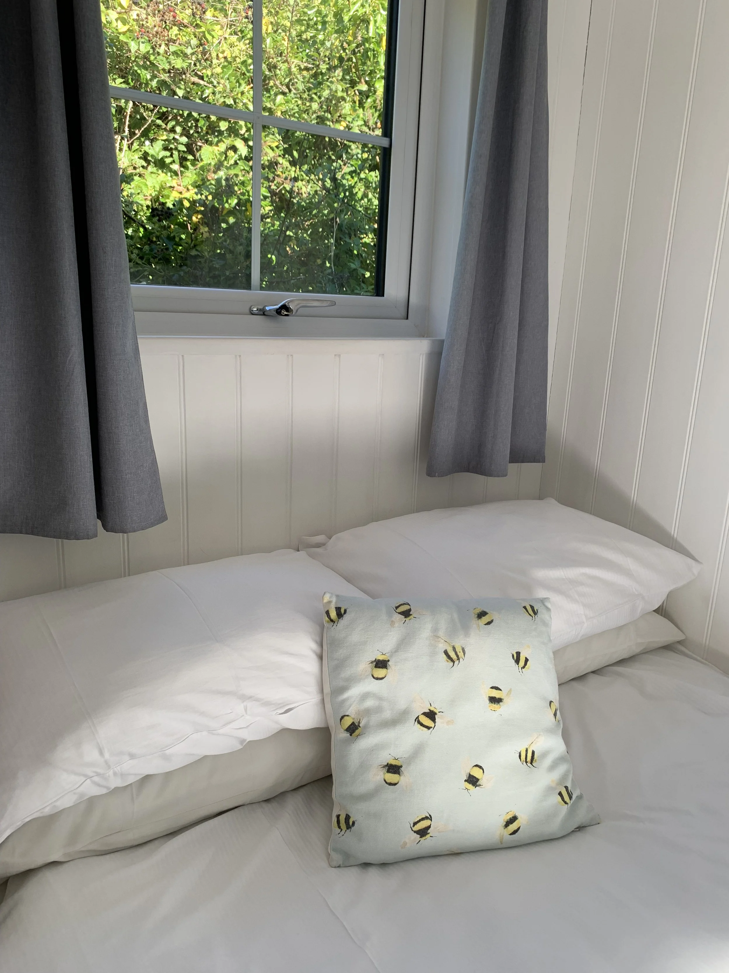 A bed with white sheets and pillows next to a window with gray curtains, and a pillow with a bee pattern. Outside the window, green foliage is visible.