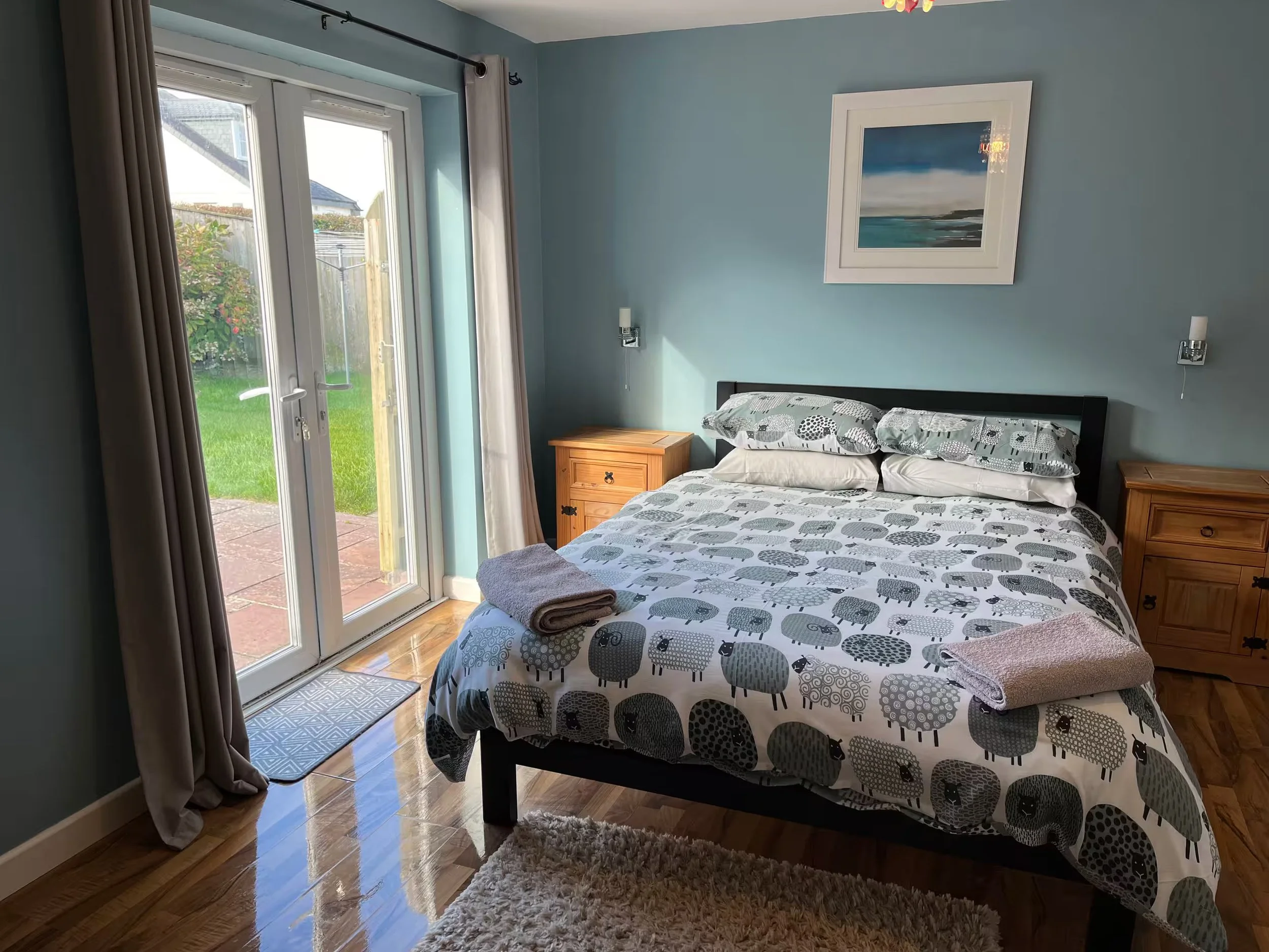 A bedroom with a double bed covered with a patterned duvet featuring sheep. There are two wooden nightstands, one on each side of the bed, and wall-mounted reading lights. A framed seascape picture hangs above the bed. French doors with beige curtain