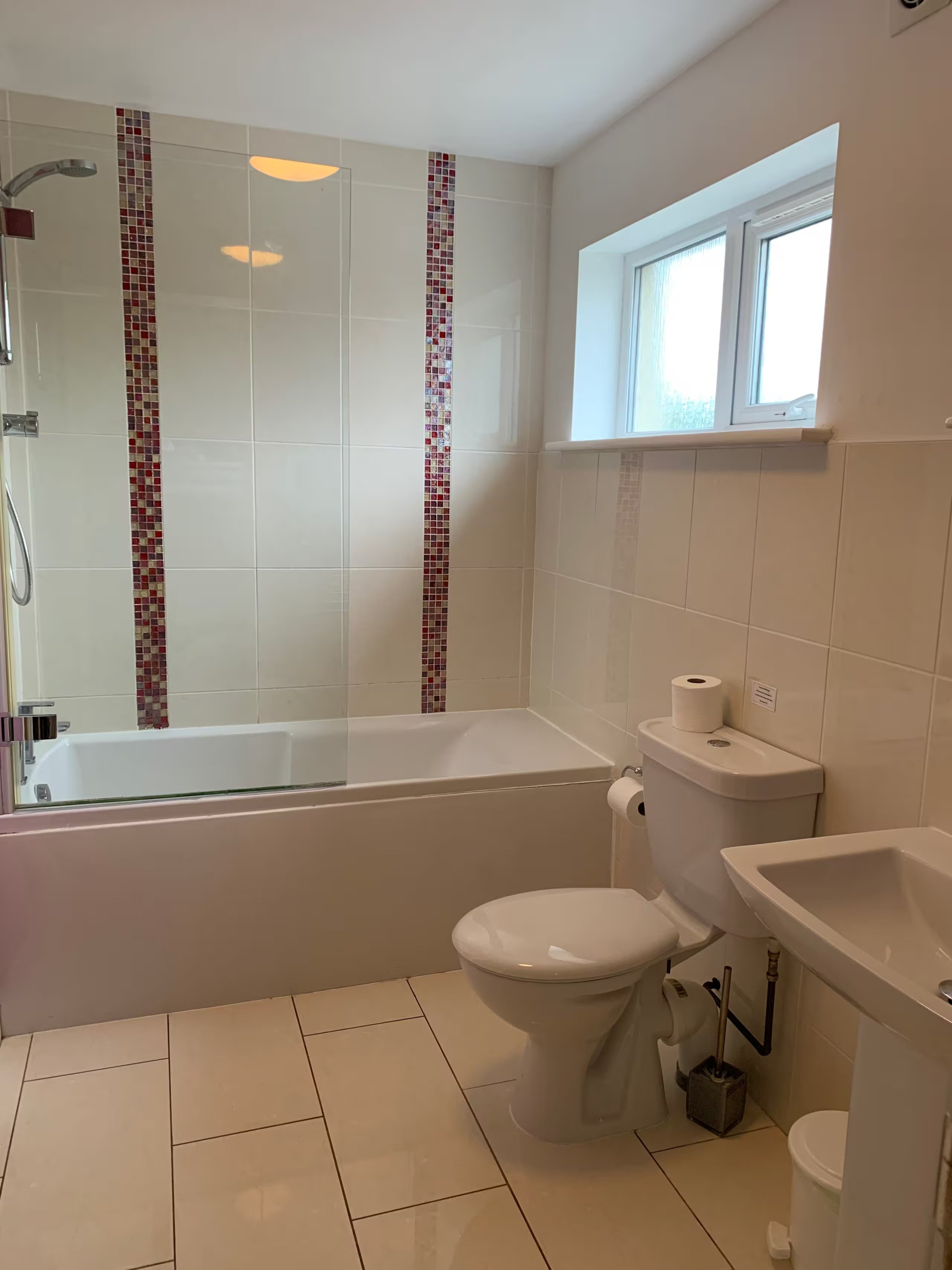 The downstairs bathroom with a bath, shower with a glass screen, a toilet and a small trash bin, a sink, a window, and beige tiled floors and walls with a red mosaic tile accent.