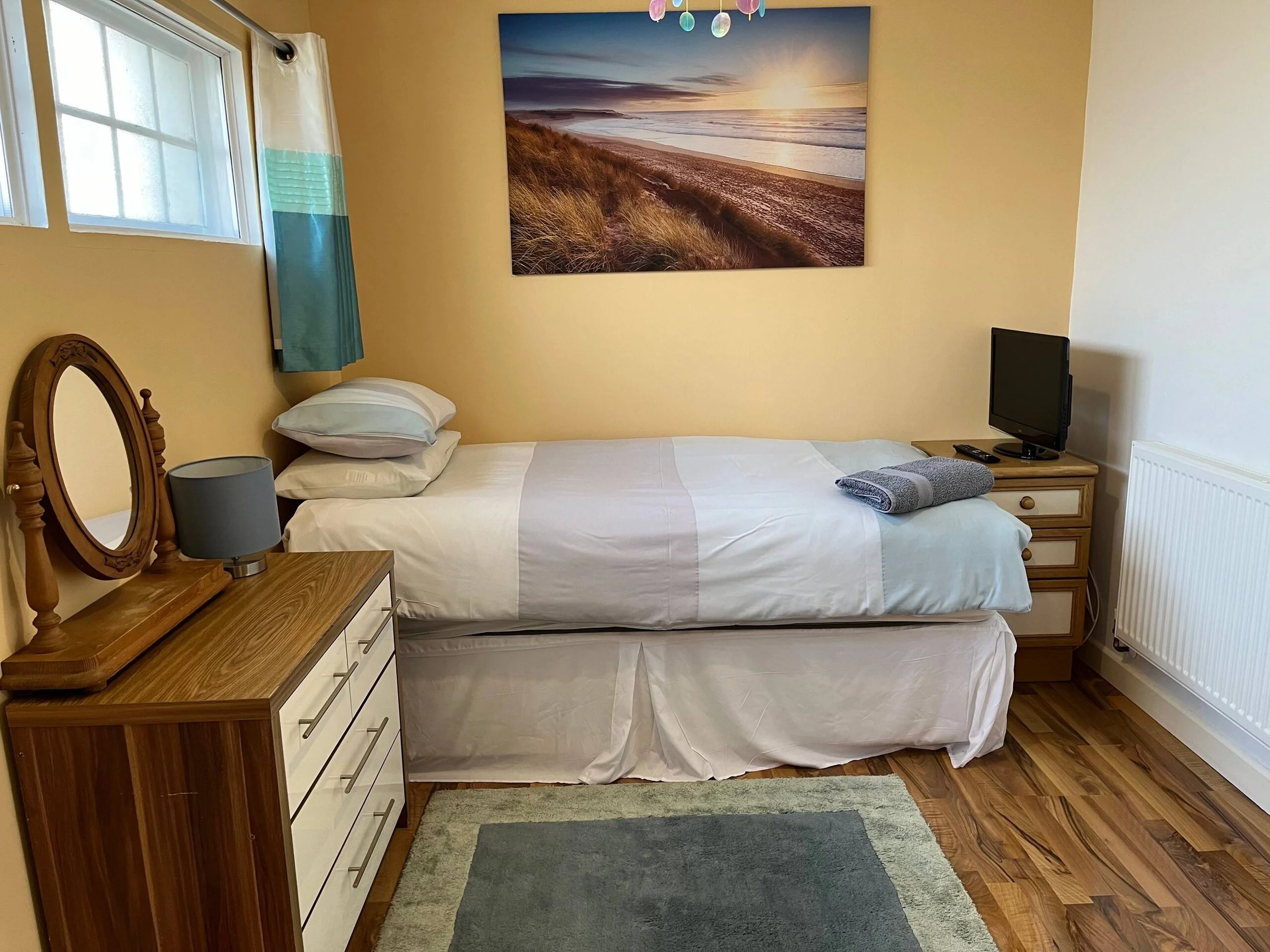 A bedroom with a bed, wooden dresser, and a small television. The wall above the bed features a large landscape photo of a beach at sunset with dunes and a sky with clouds. There is a window with light-colored curtains, and the floor is covered with 
