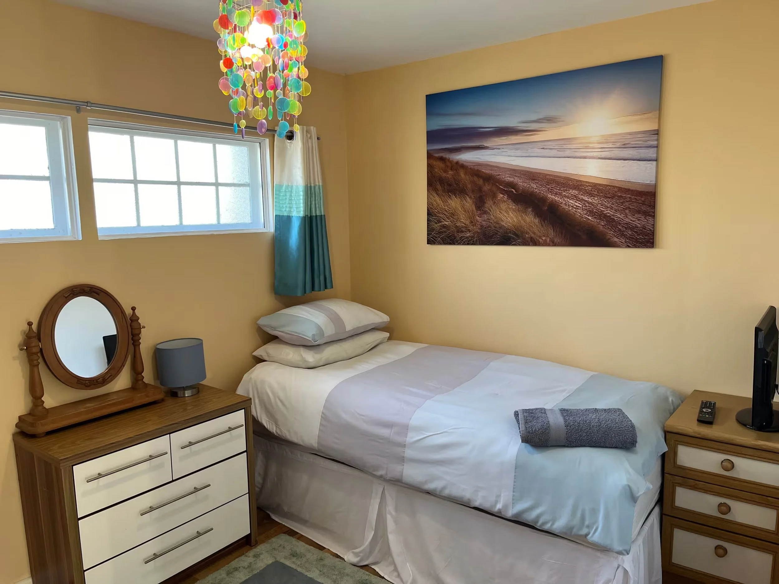 A bedroom with a bed, two nightstands with lamps and drawers, a wall art of a beach sunset, and a colorful hanging light fixture.