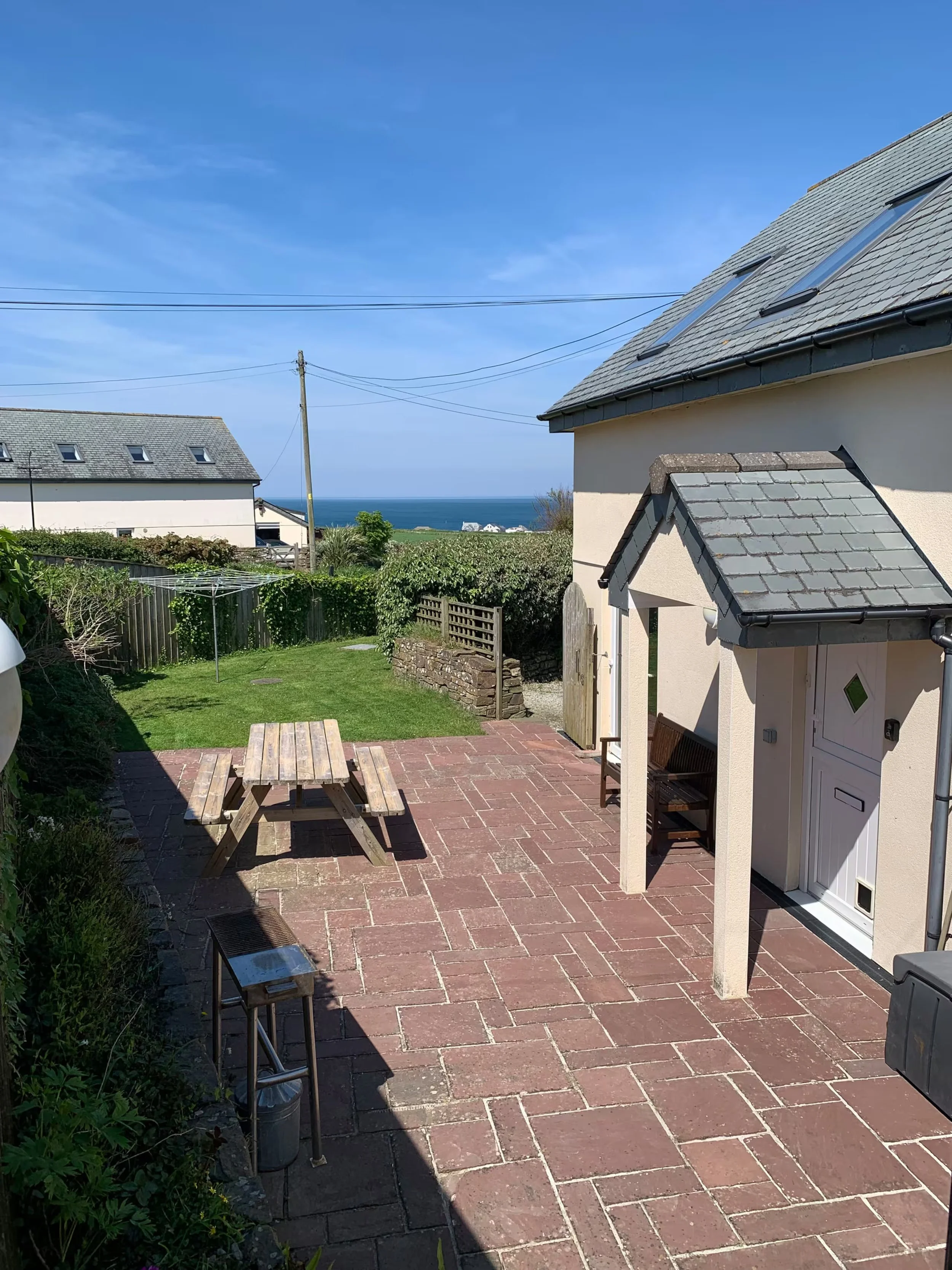 An upper patio looking at  a wooden picnic table and garden with greenery. A view of the ocean in the background under a blue sky.