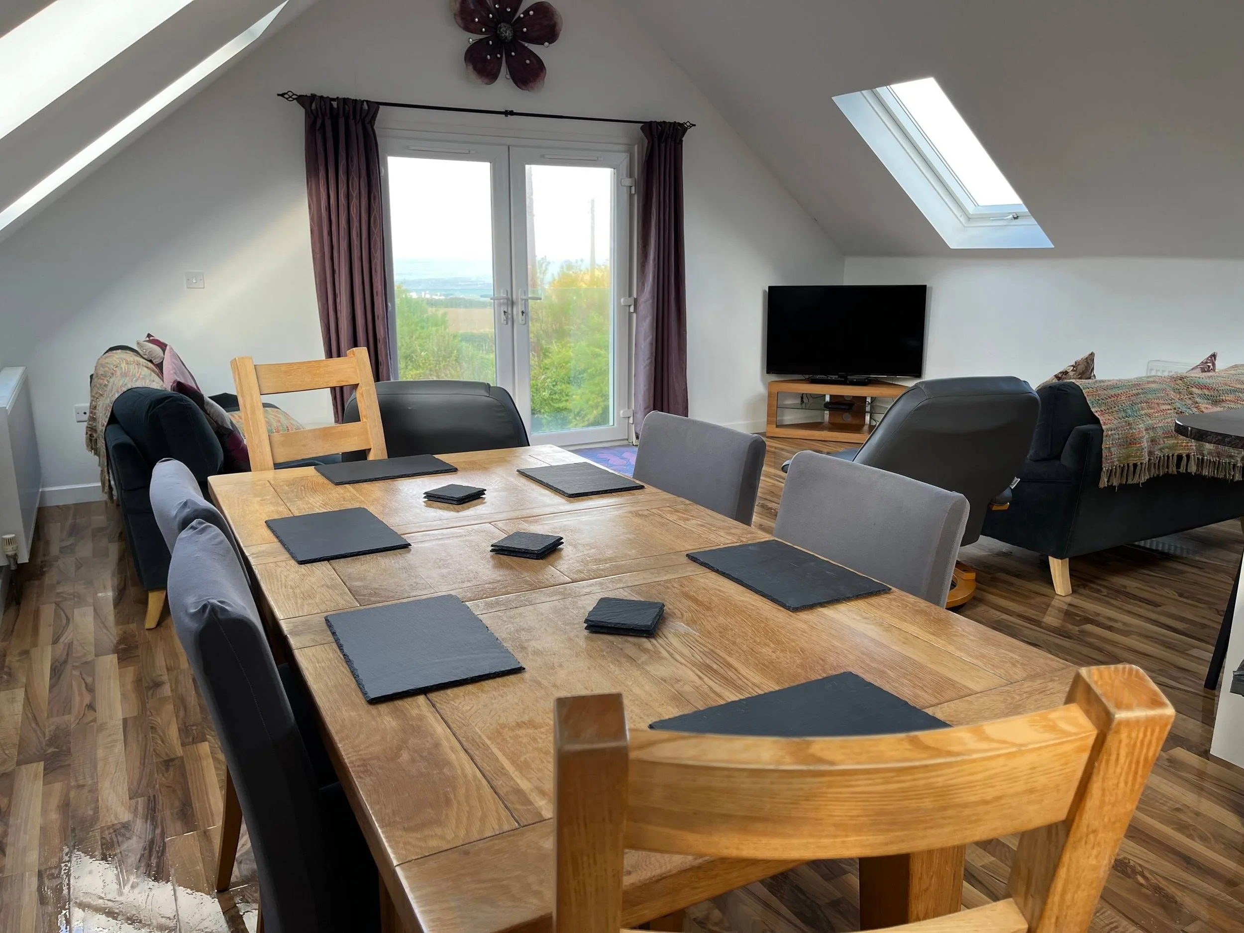 A dining area with a large wooden table set with black placemats, surrounded by a mix of padded and wooden chairs. There is a windowed door with purple curtains, a sofa with patterned and solid cushions, and a television on a wooden stand. Large skyl