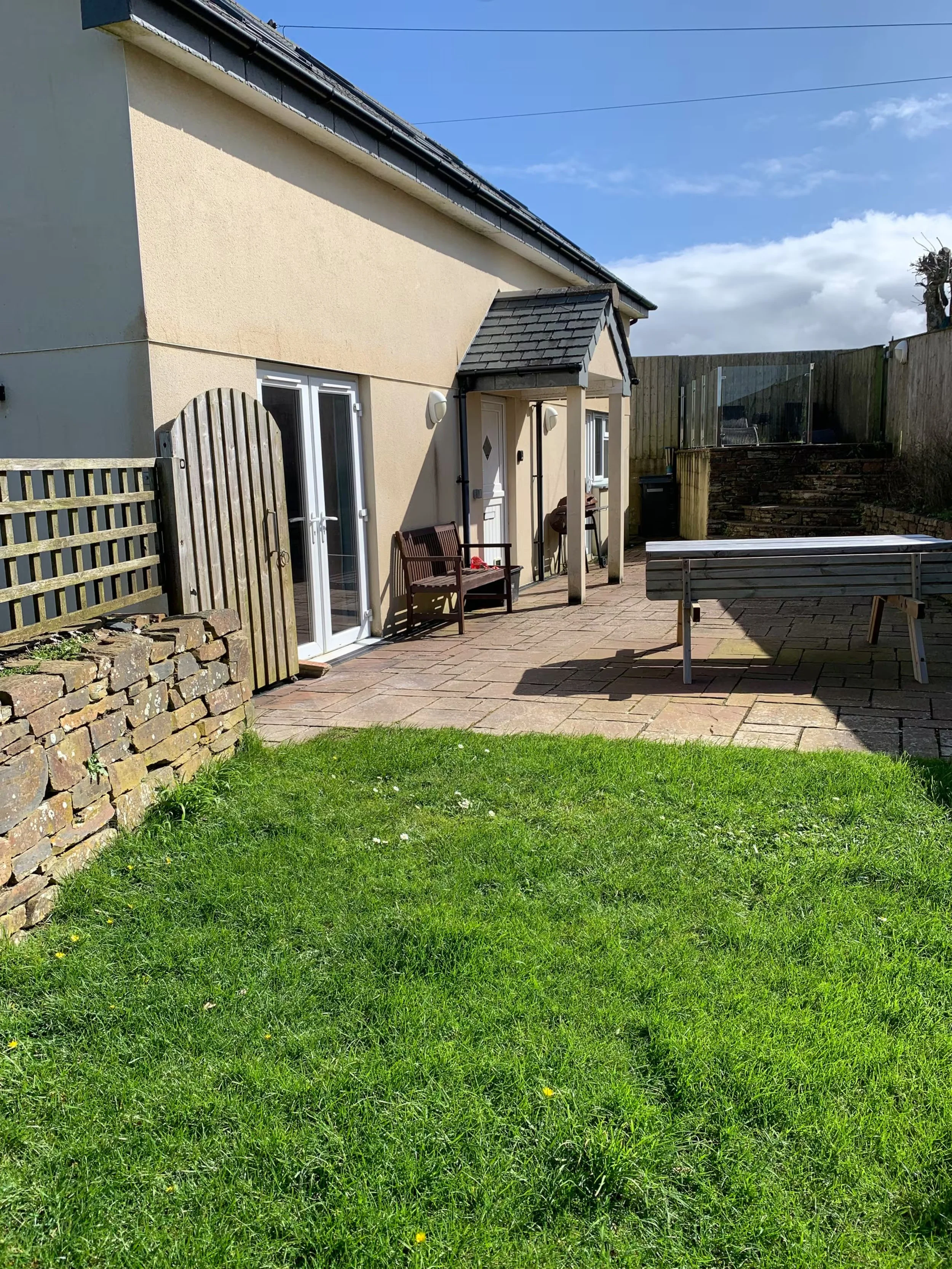 Backgarden at Sunnyridge with a grassy lawn, a wooden fence, a stone border, patio area with benches, and part of a beige house with patio glass doors and a small porch.