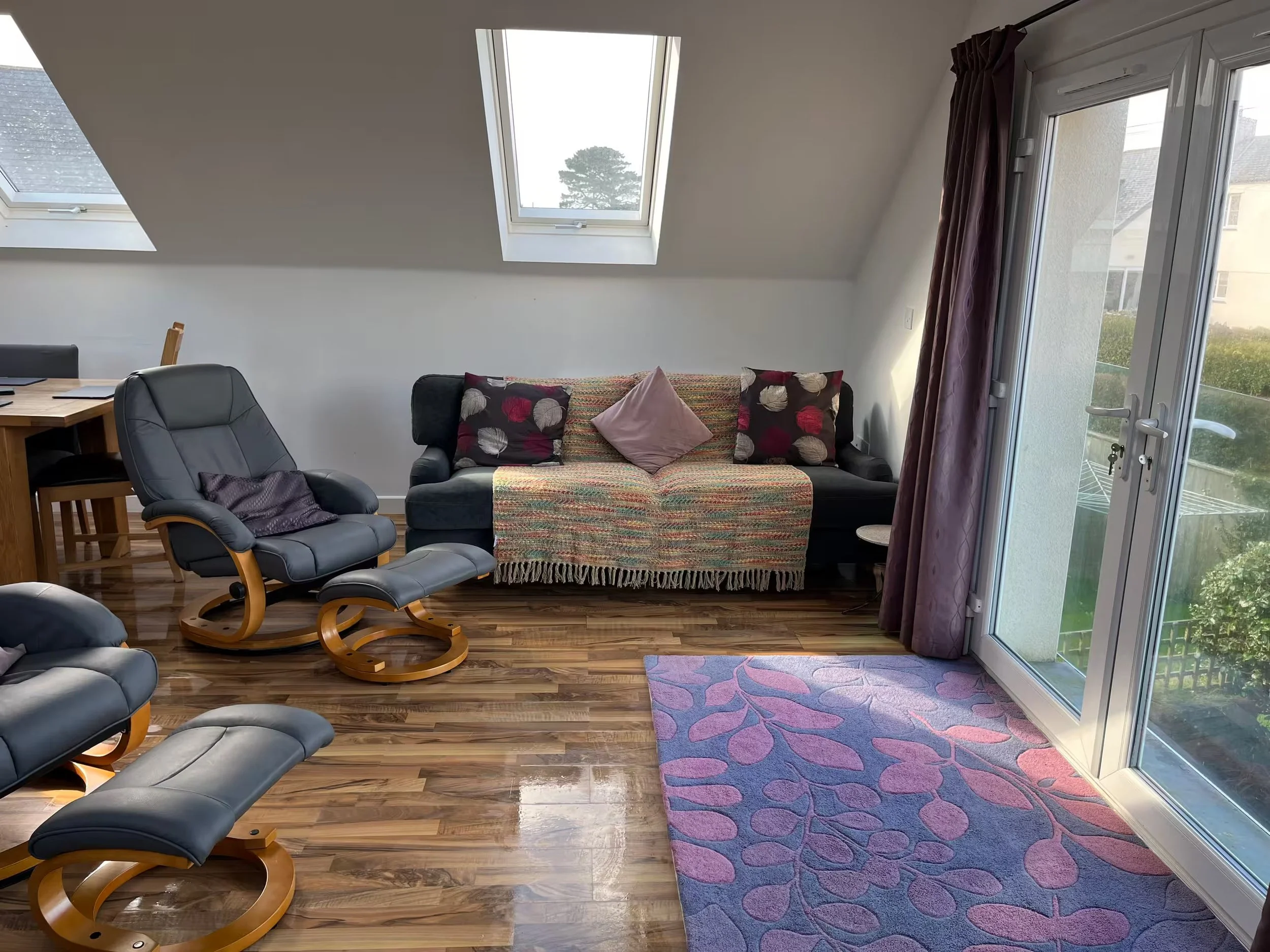 Sunnyridge living room with sloped ceiling, skylights, a couch with pillows, a purple curtain, and glass doors leading to a balcony with a view of trees and neighboring buildings.