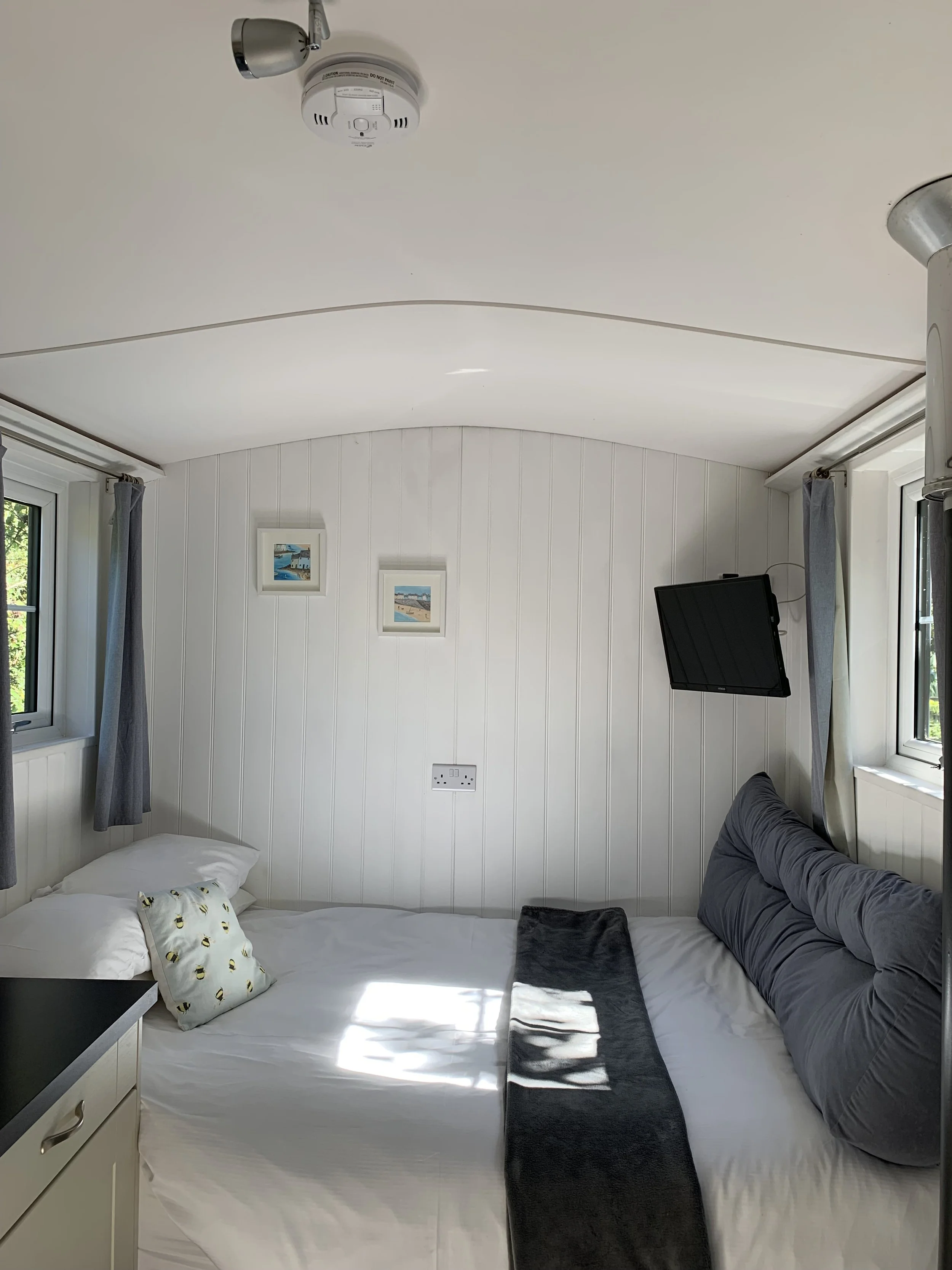 A cozy bedroom with white panel walls, two windows with gray curtains, a bed with white bedding, a gray pillow, a black throw blanket, a pillow with bee patterns, a small black table, a wall-mounted TV, three small framed pictures of beach scenes, an