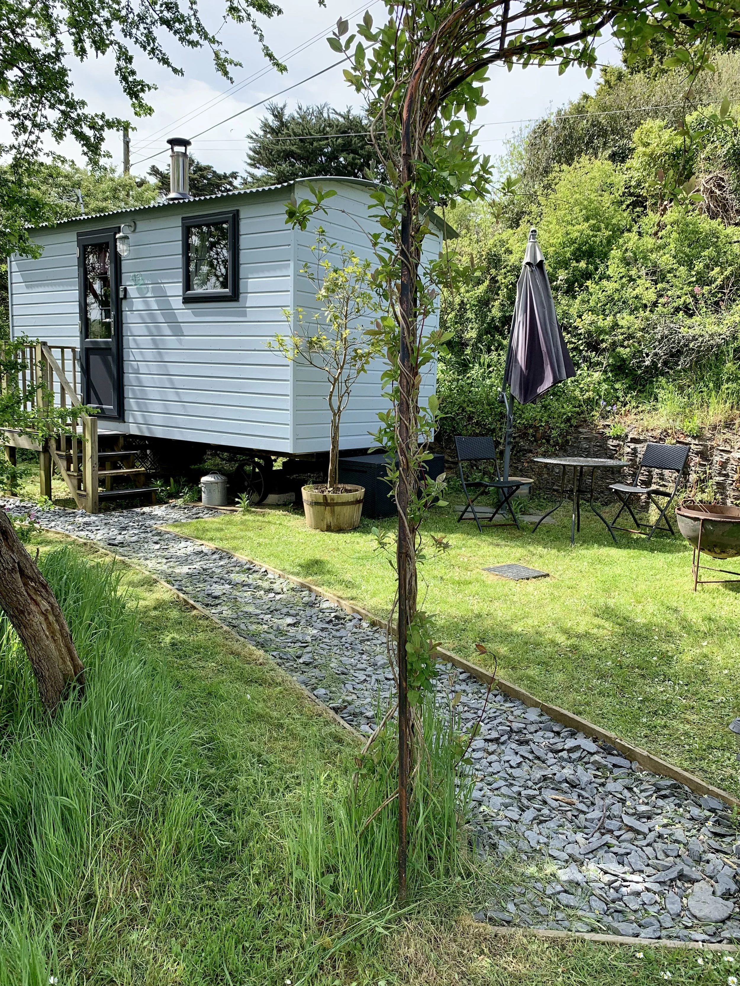 A small white mobile home on a grassy yard with trees, a rock pathway, outdoor furniture, and a closed umbrella.