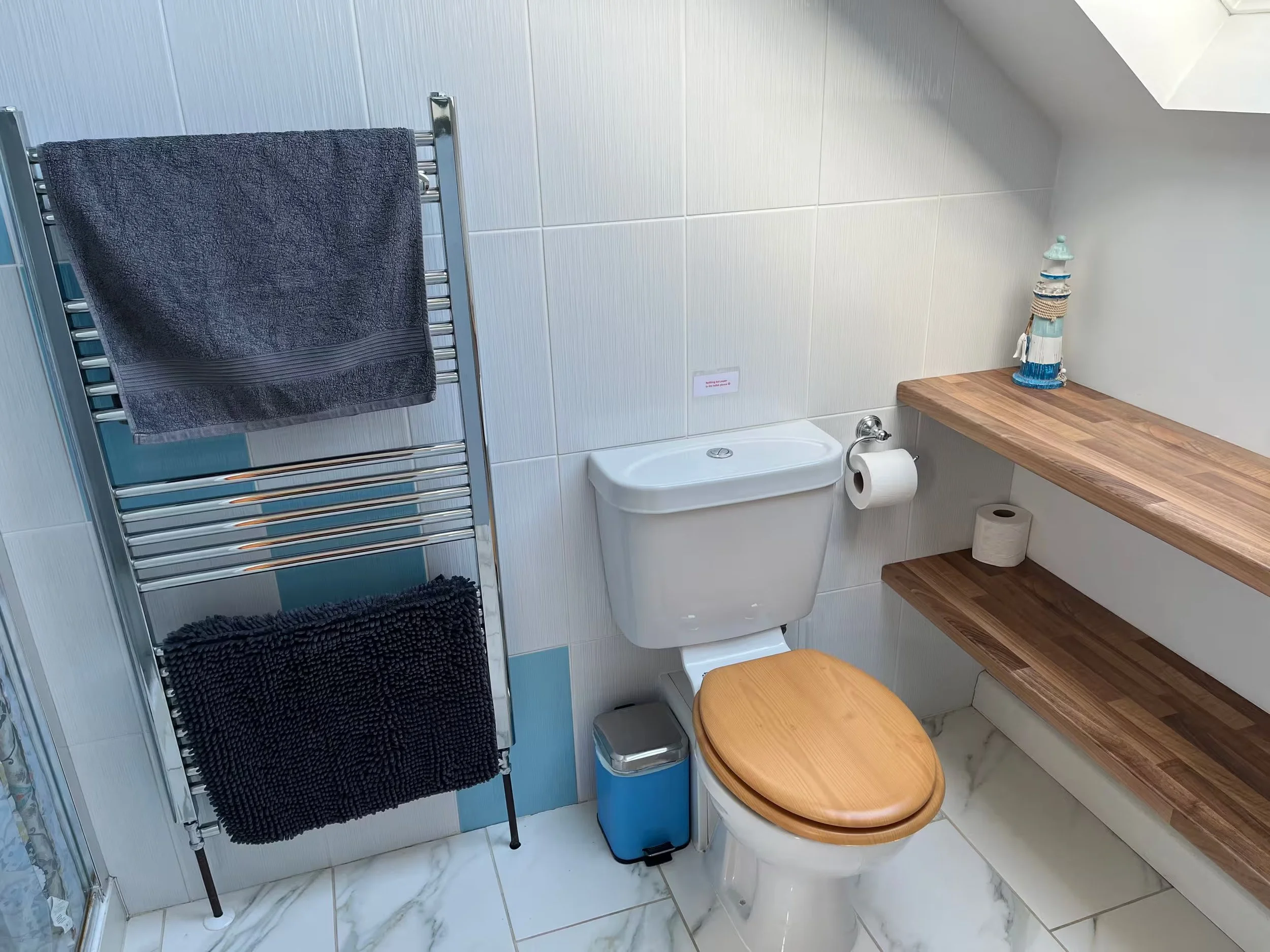 Upstairs bathroom with white tiled walls, a toilet with a wooden seat, a wooden countertop, and a decorative lighthouse on the countertop. There is a towel rail with dark towels and a small trash bin.