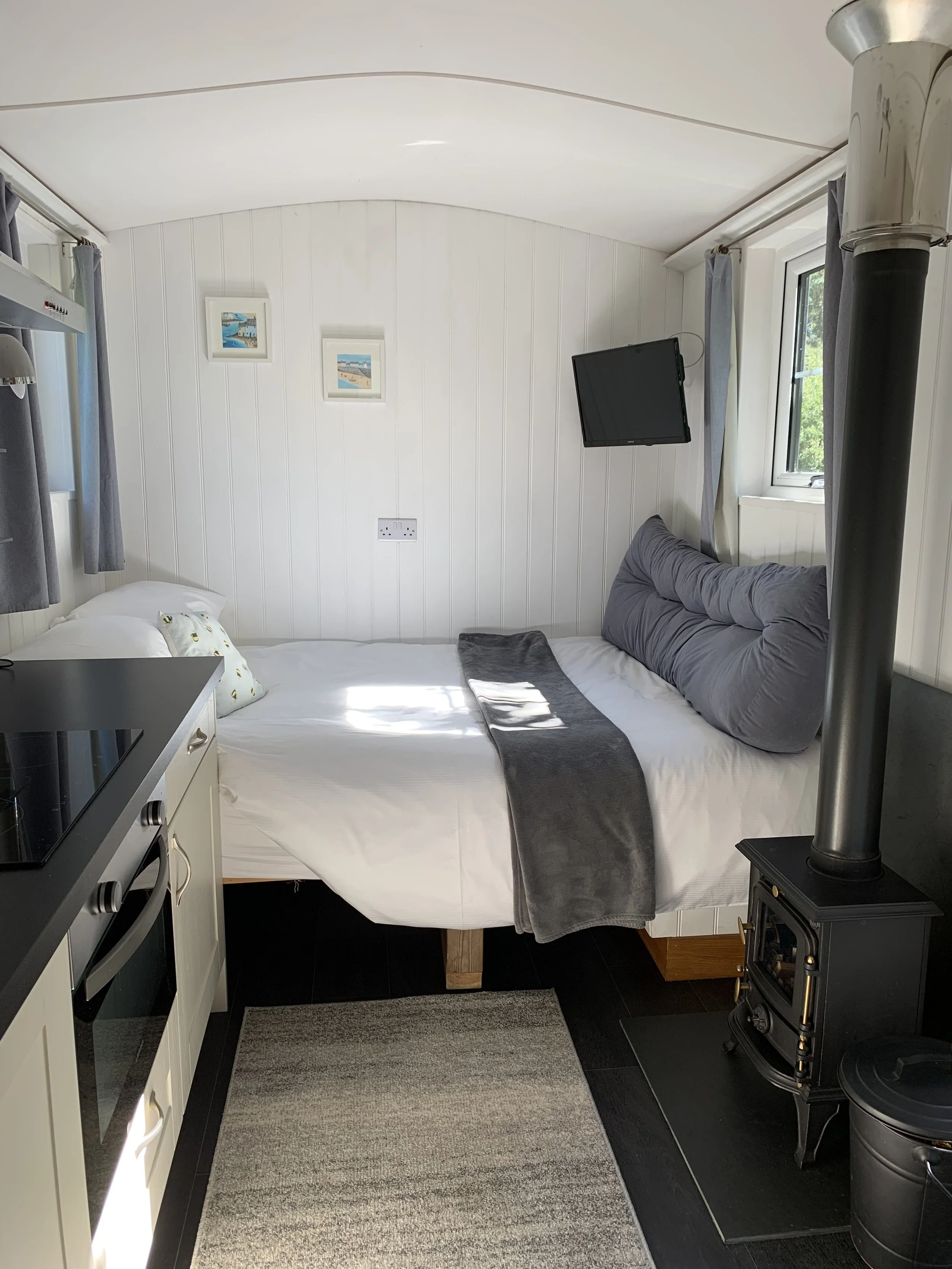 A cozy living space with a bed and a wood stove. The bed has white sheets, pillows, and a gray blanket, with a gray and a decorative pillow. A small rug is on the floor beneath the bed. There's a window with curtains, and a flat-screen TV mounted on 