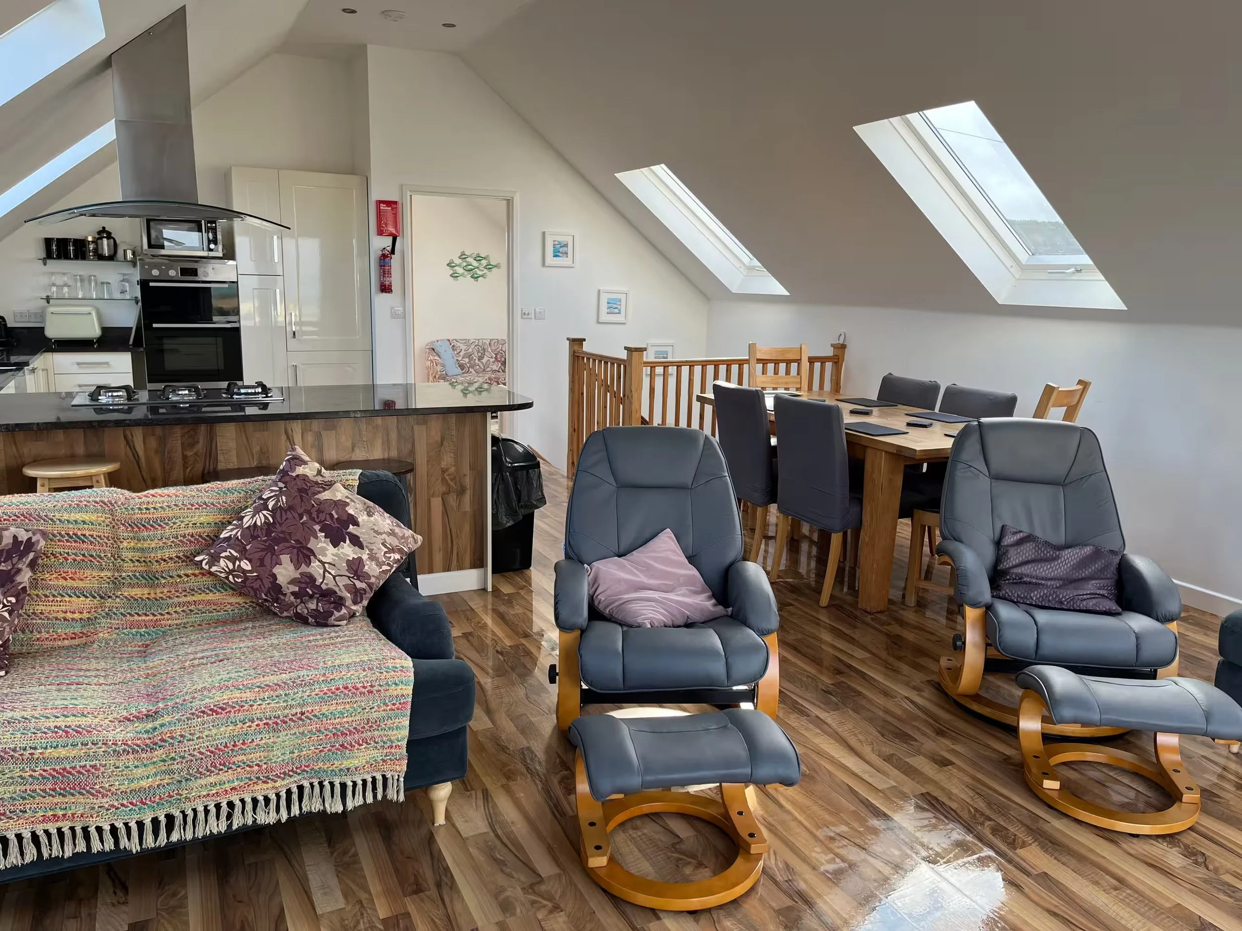 Spacious reverse accommodation at Sunnyridge with kitchen, dining area, several black leather rocking chairs with wooden frames, a colourful blanket and pillows on a sofa, and skylight windows in the ceiling.