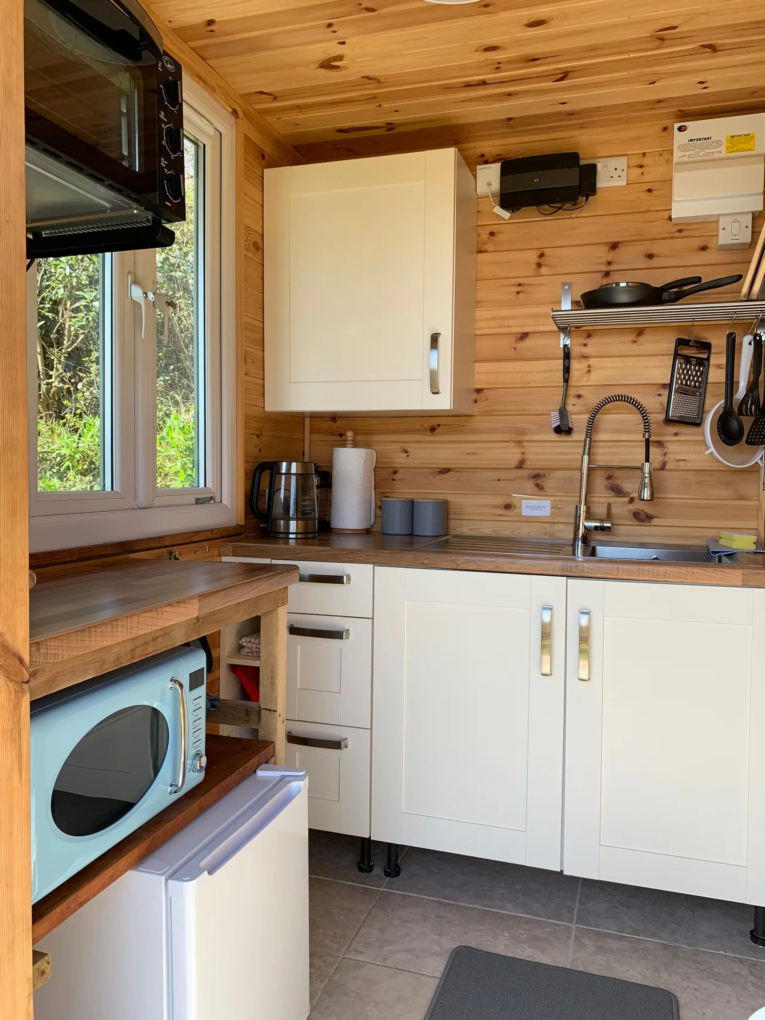 A cozy kitchen at Foxes Cove with wood-paneled walls and ceiling, a window overlooking greenery, white cabinetry, a sink with a modern faucet, and various kitchen appliances including a microwave, kettle, and a small oven.