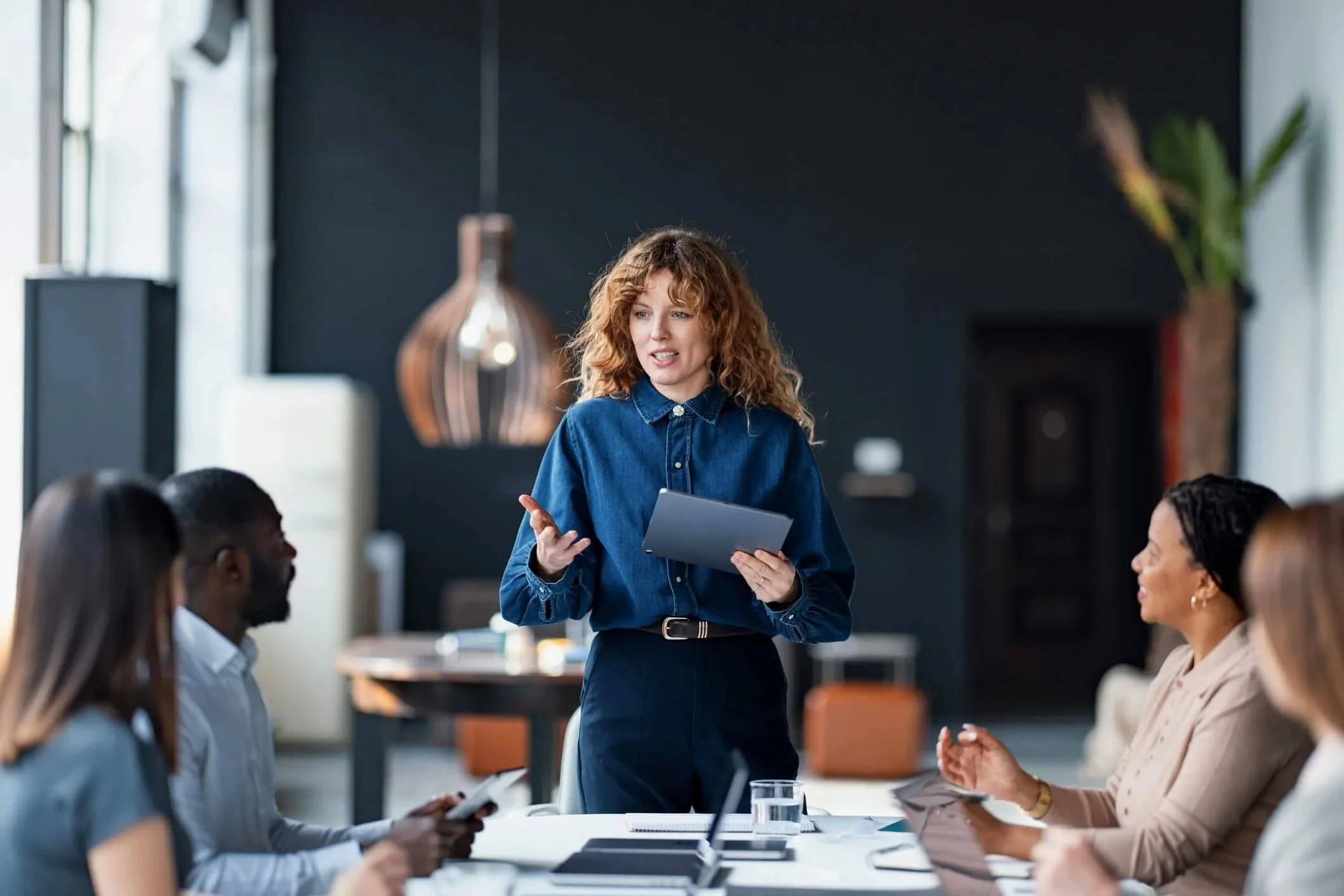 A woman in a denim shirt speaking to a group of diverse people during a meeting in a modern office.