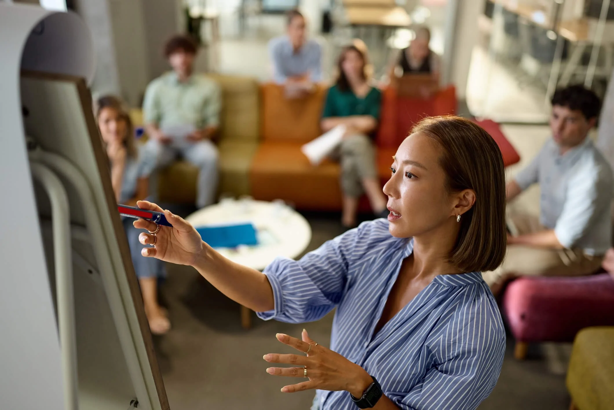 A woman with short hair and earrings using a stylus on a touchscreen device in a modern, well-lit office space with blurred people sitting on couches in the background.