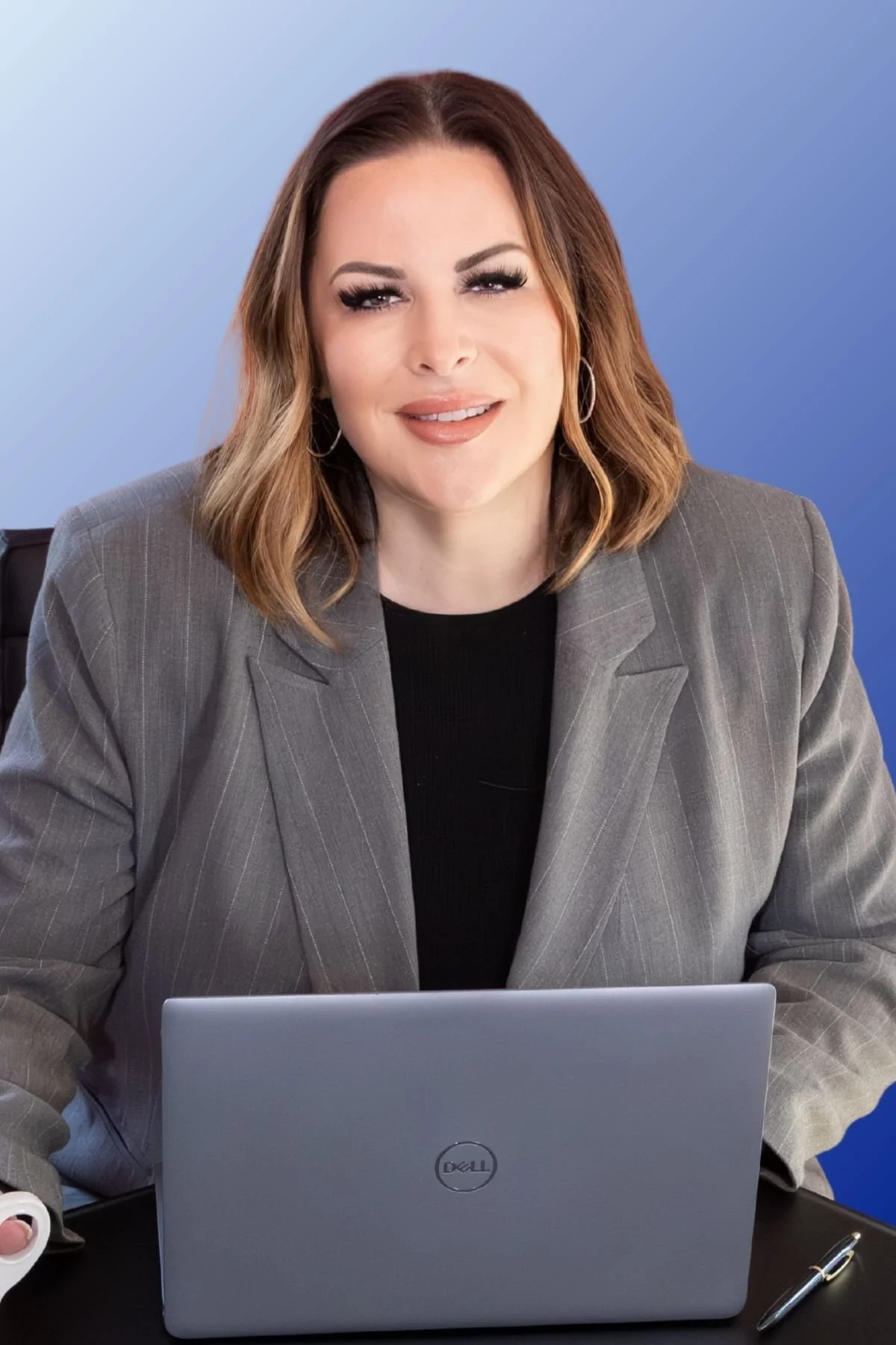 A woman with shoulder-length hair, wearing a gray blazer and black top, sitting at a desk with a silver Dell laptop in front of her against a blue gradient background.