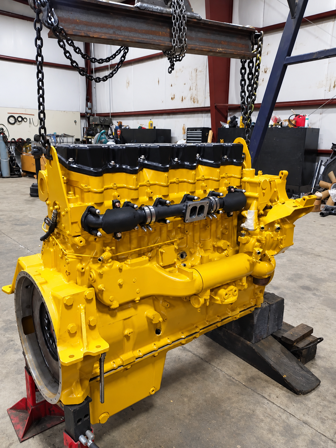 Yellow diesel engine with black valve covers beneath a chain hoist in a workshop.