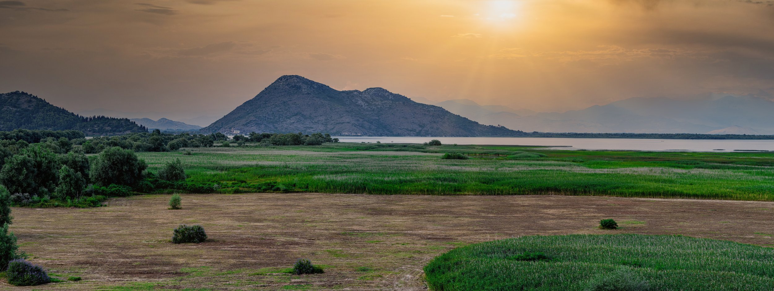 Scenic landscape with green fields, trees, mountains in the distance, and a body of water, under a golden sky at sunset.