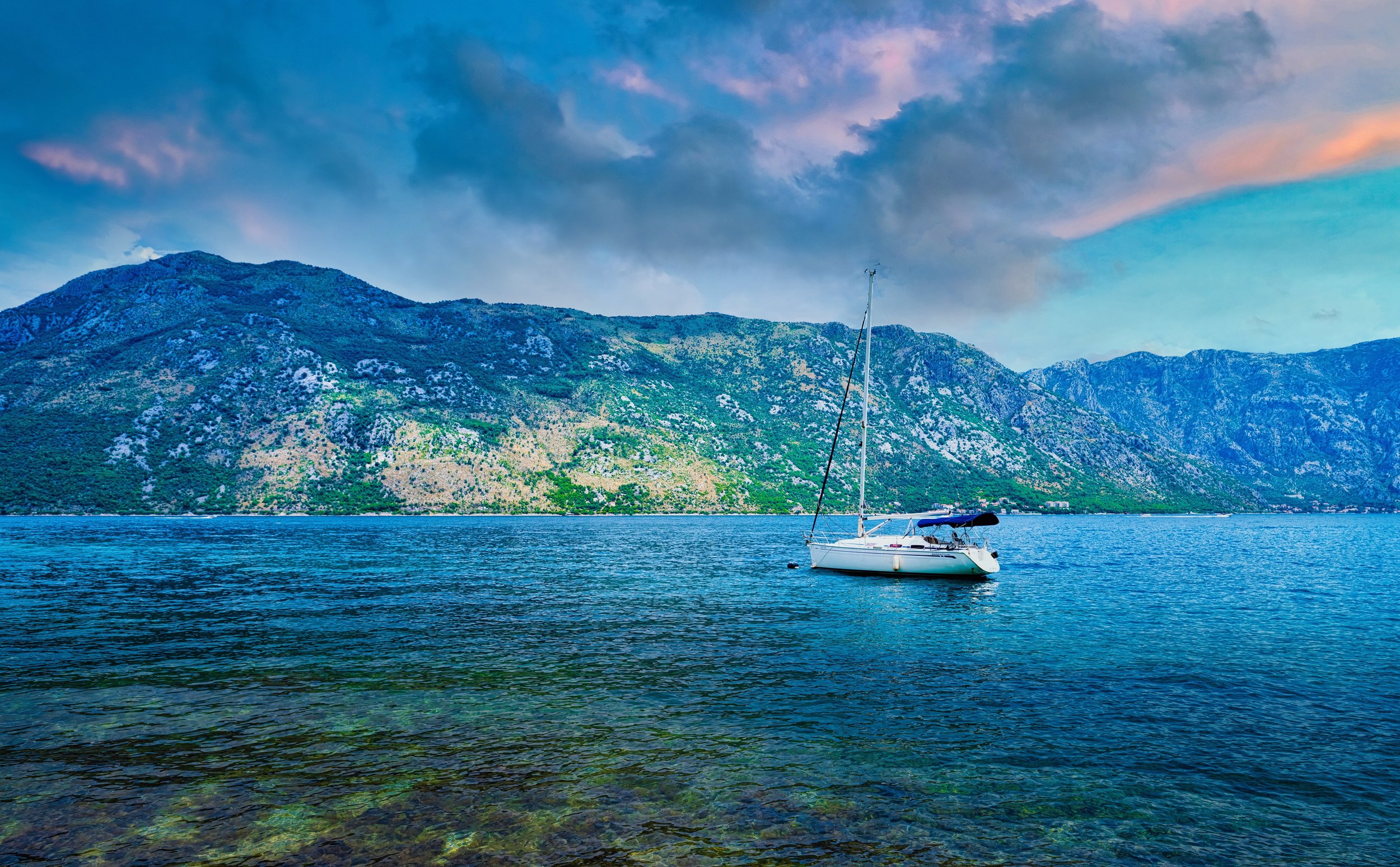A sailboat floating on calm water with a mountainous landscape and pink and blue sky in the background.