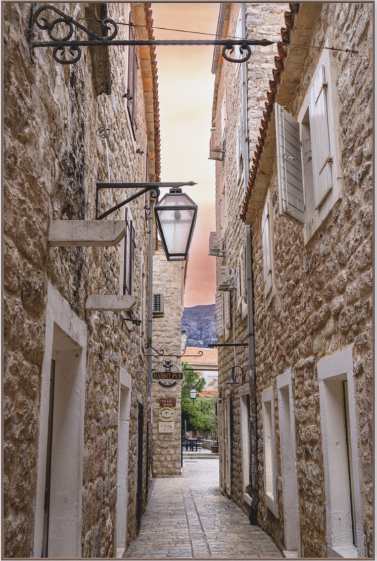 Narrow cobblestone alley enclosed by old stone buildings with white shutters and vintage street lamps, leading to a small courtyard with outdoor seating and a mountainous landscape in the background.