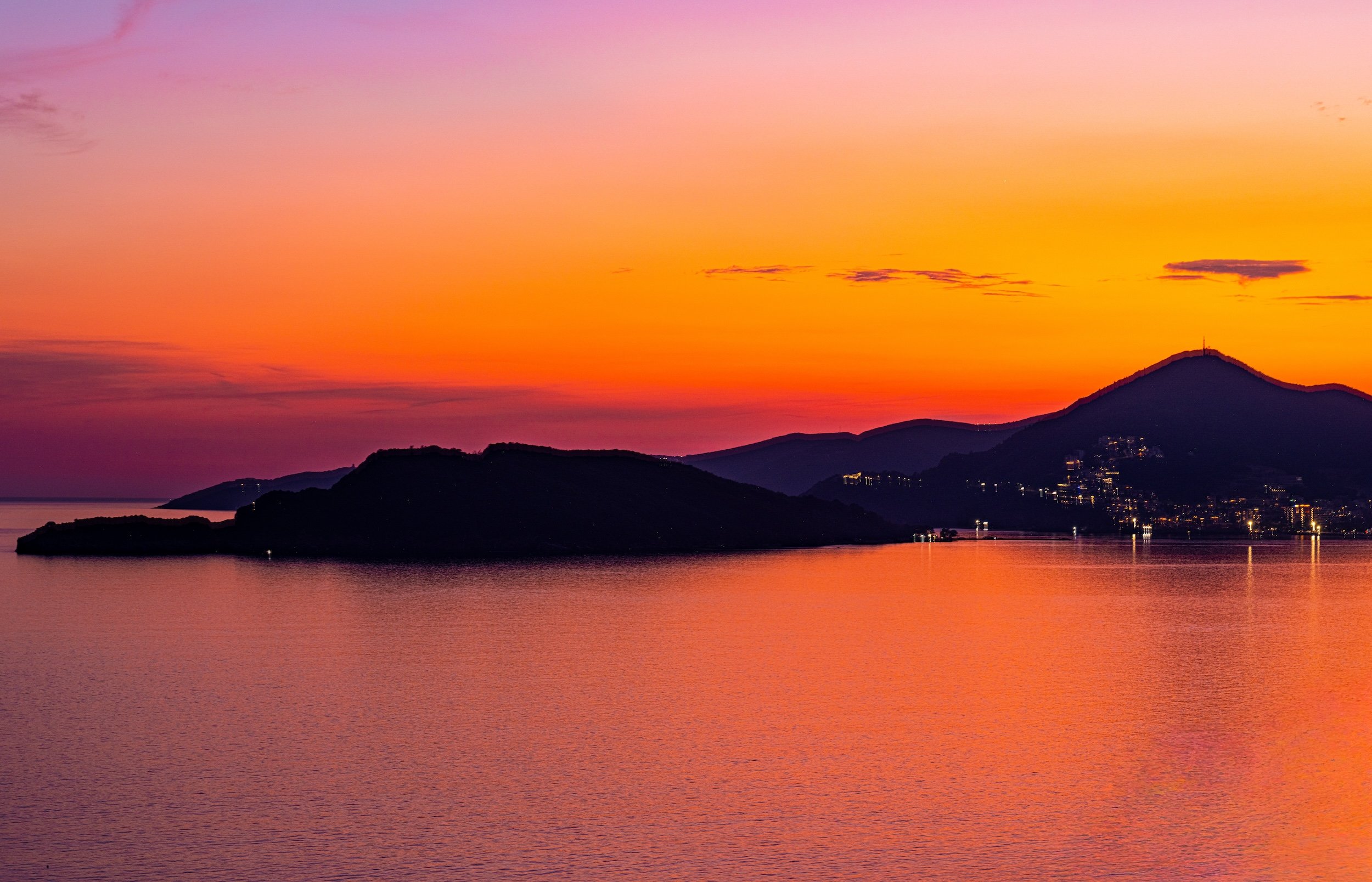 Sunset over a body of water with silhouettes of hills and mountains, some with illuminated buildings, featuring a colorful gradient sky with shades of pink, orange, and purple.