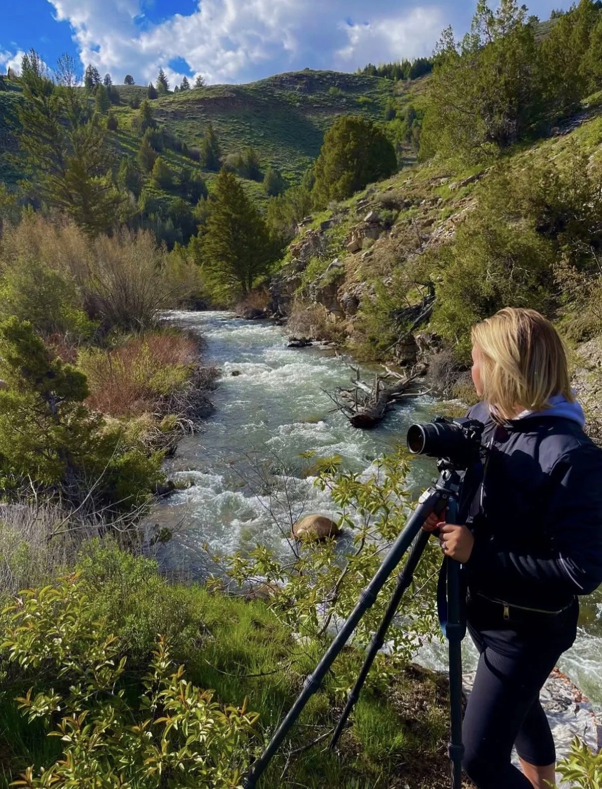 Woman with blonde hair holding a telescope by a river in a forested mountainous area.