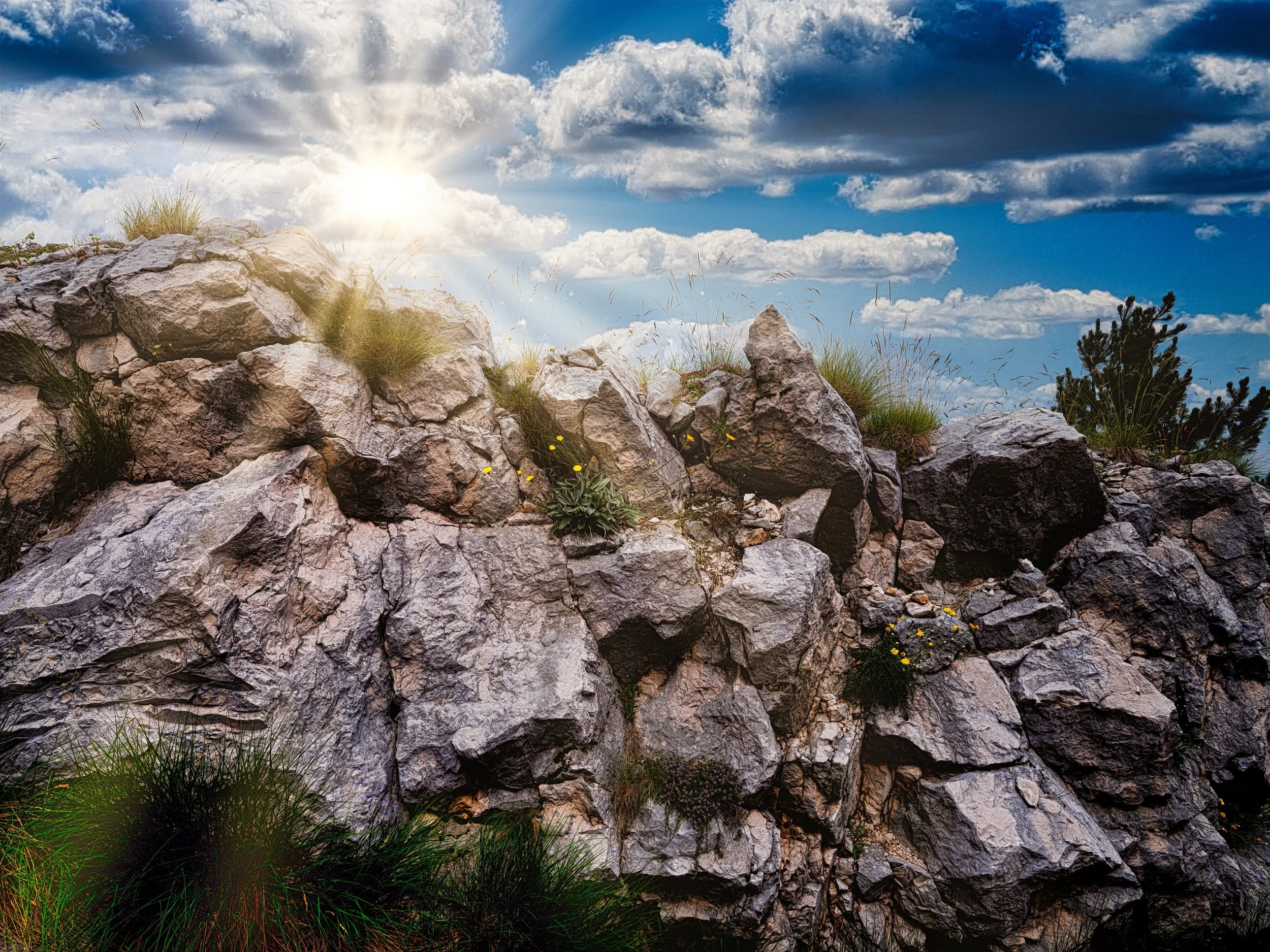 A rocky hillside with patches of grass and small plants, under a partly cloudy blue sky with the sun shining through clouds.
