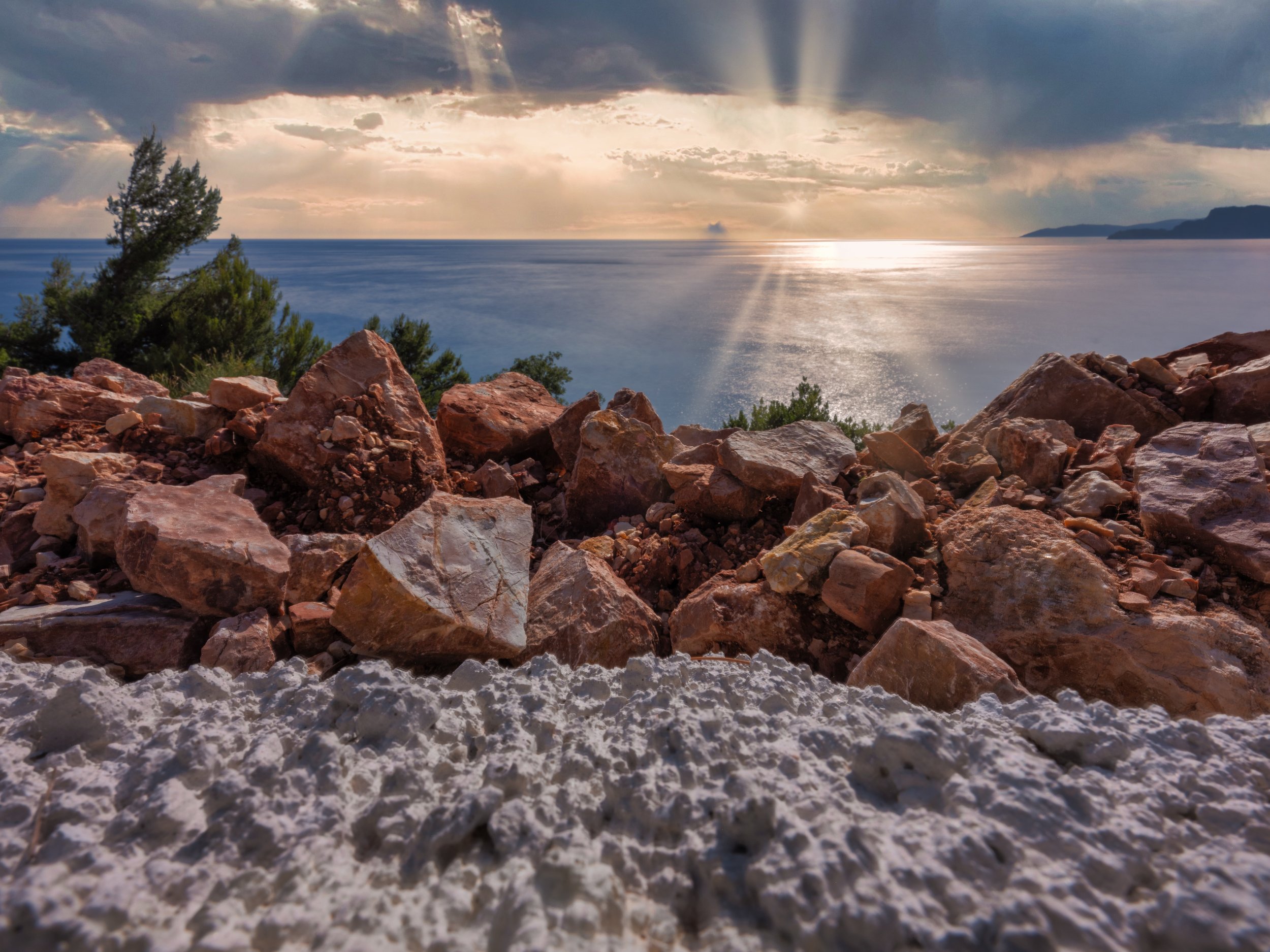 View of rocky shore with a body of water and distant mountains under a sky with dark clouds and sunlight breaking through.