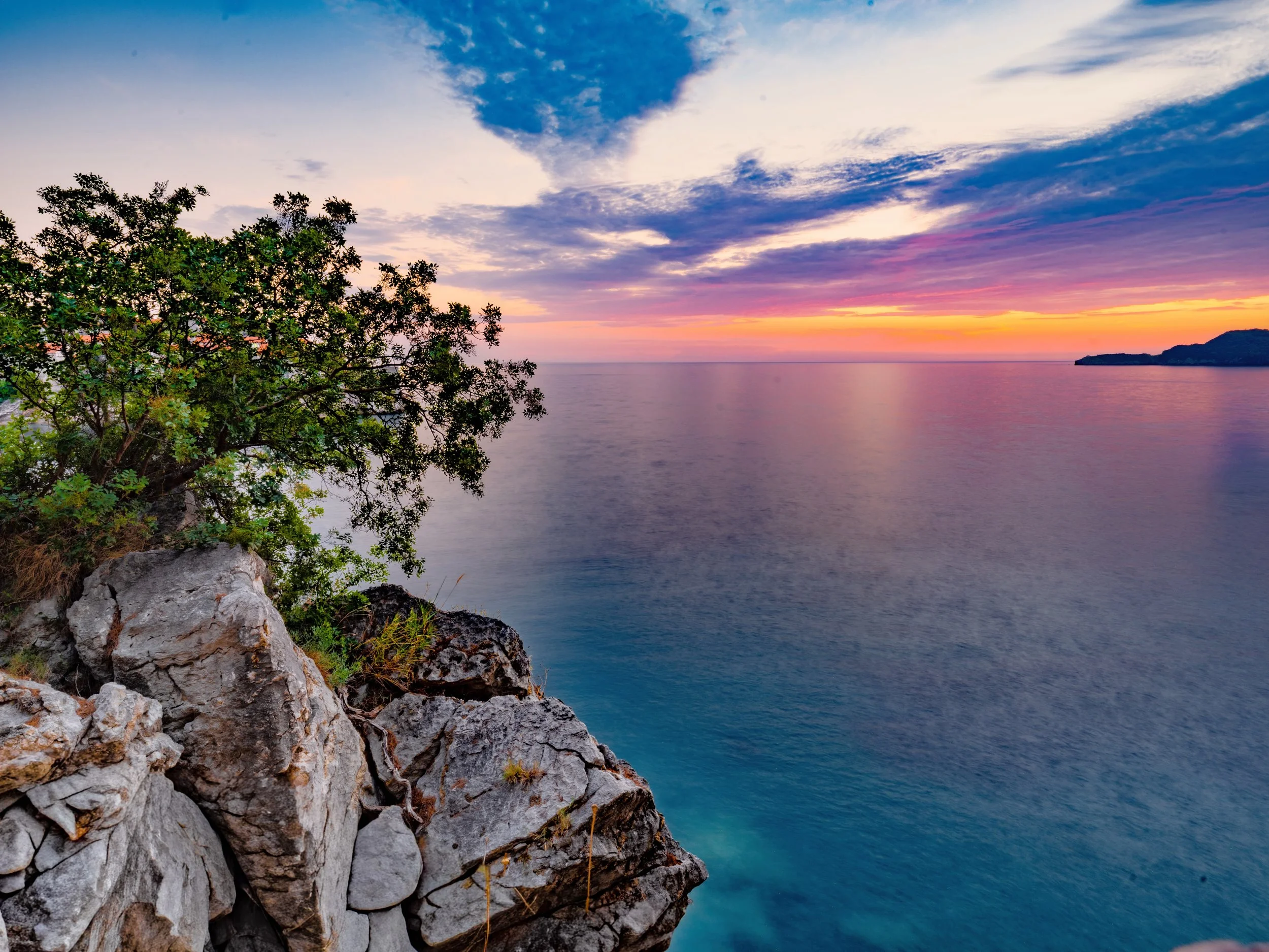 Sunset over a body of water with colorful sky, trees, and rocky shoreline in the foreground.