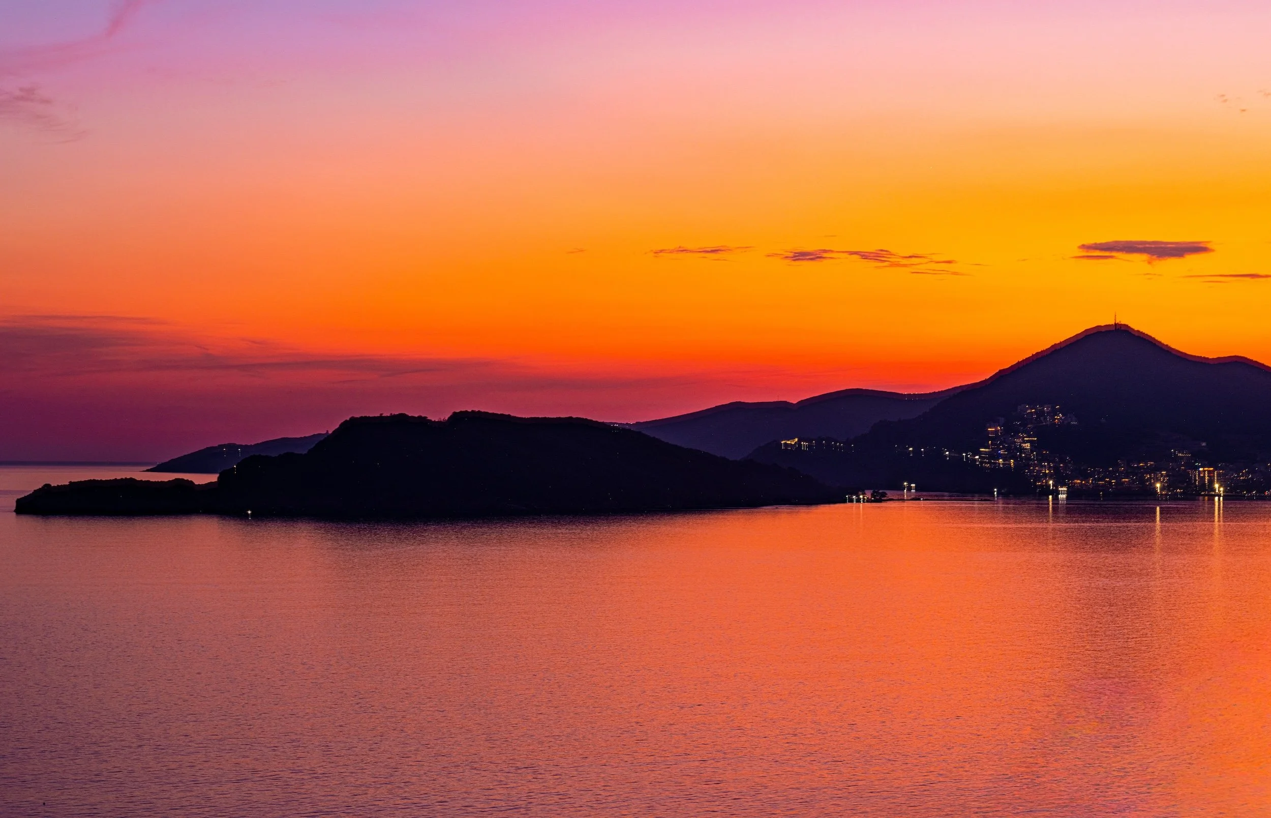 Sunset over the mountains with a body of water in the foreground, displaying shades of pink, orange, and purple in the sky.