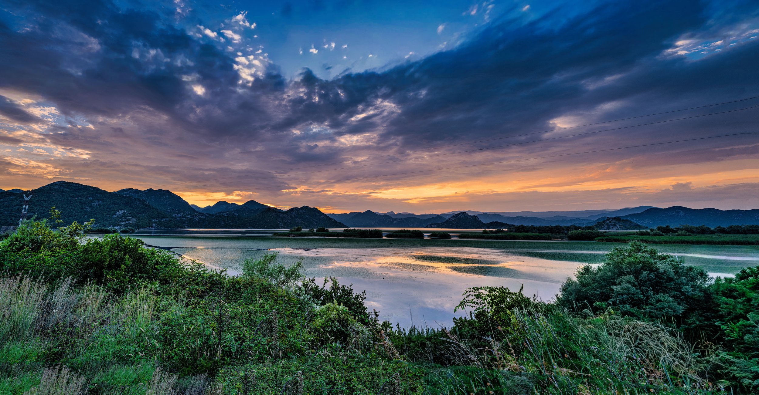 A scenic view of a river with greenery in the foreground, mountains in the background, and a sky with scattered clouds at sunset.