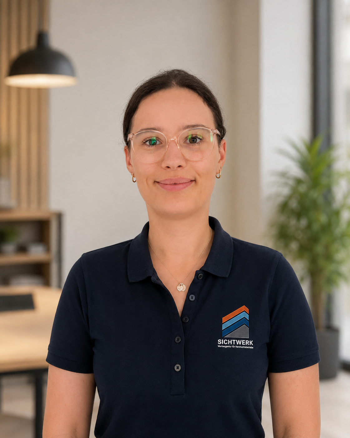 Junge Frau trägt eine dunkelblaue Poloshirt mit dem Logo von SICHTWERK, steht in einem modern eingerichteten Raum mit Pflanzen und Regalen im Hintergrund.