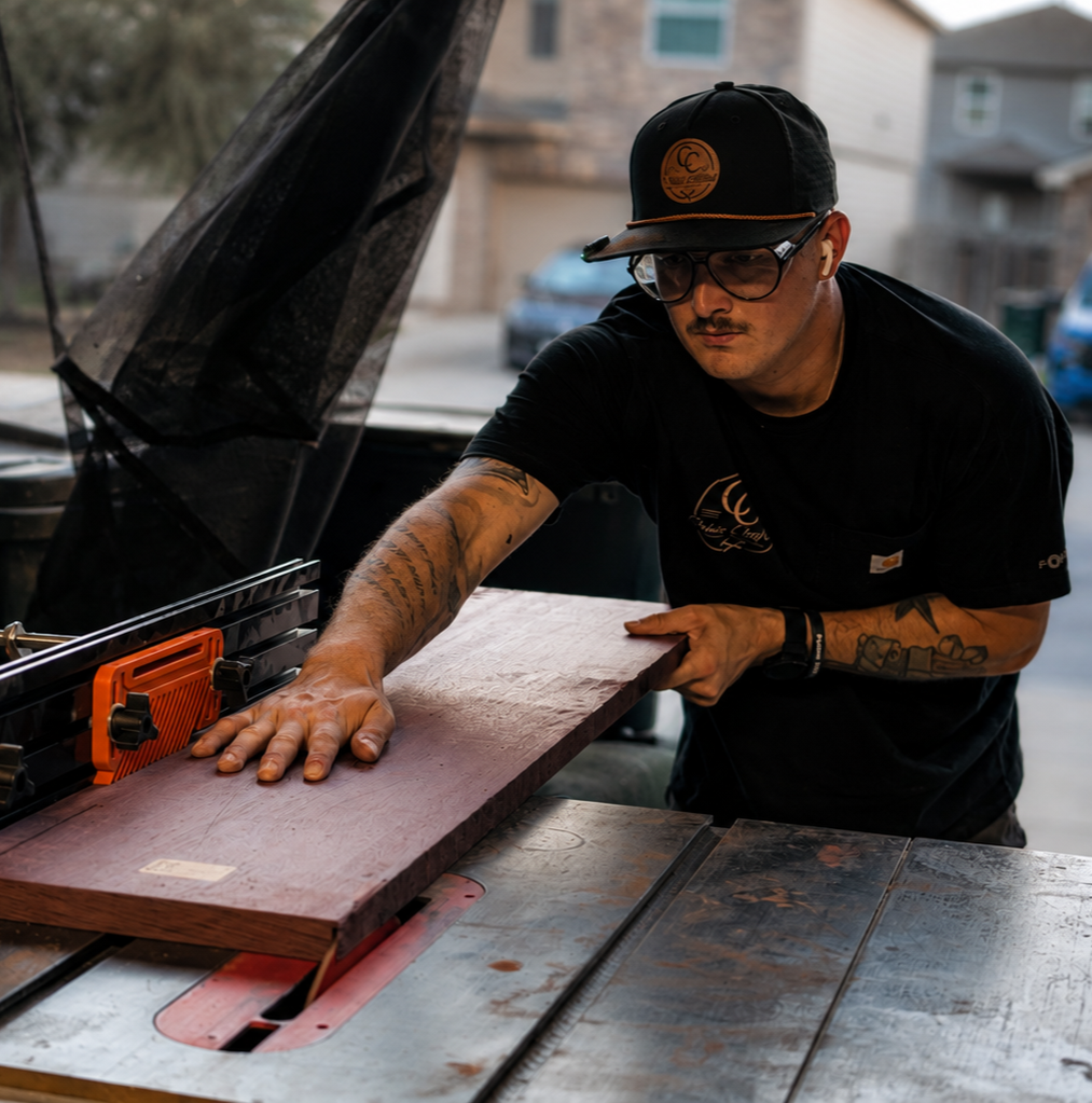 A man with tattoos, glasses, and a black cap is cutting a piece of wood with a circular saw outdoors.