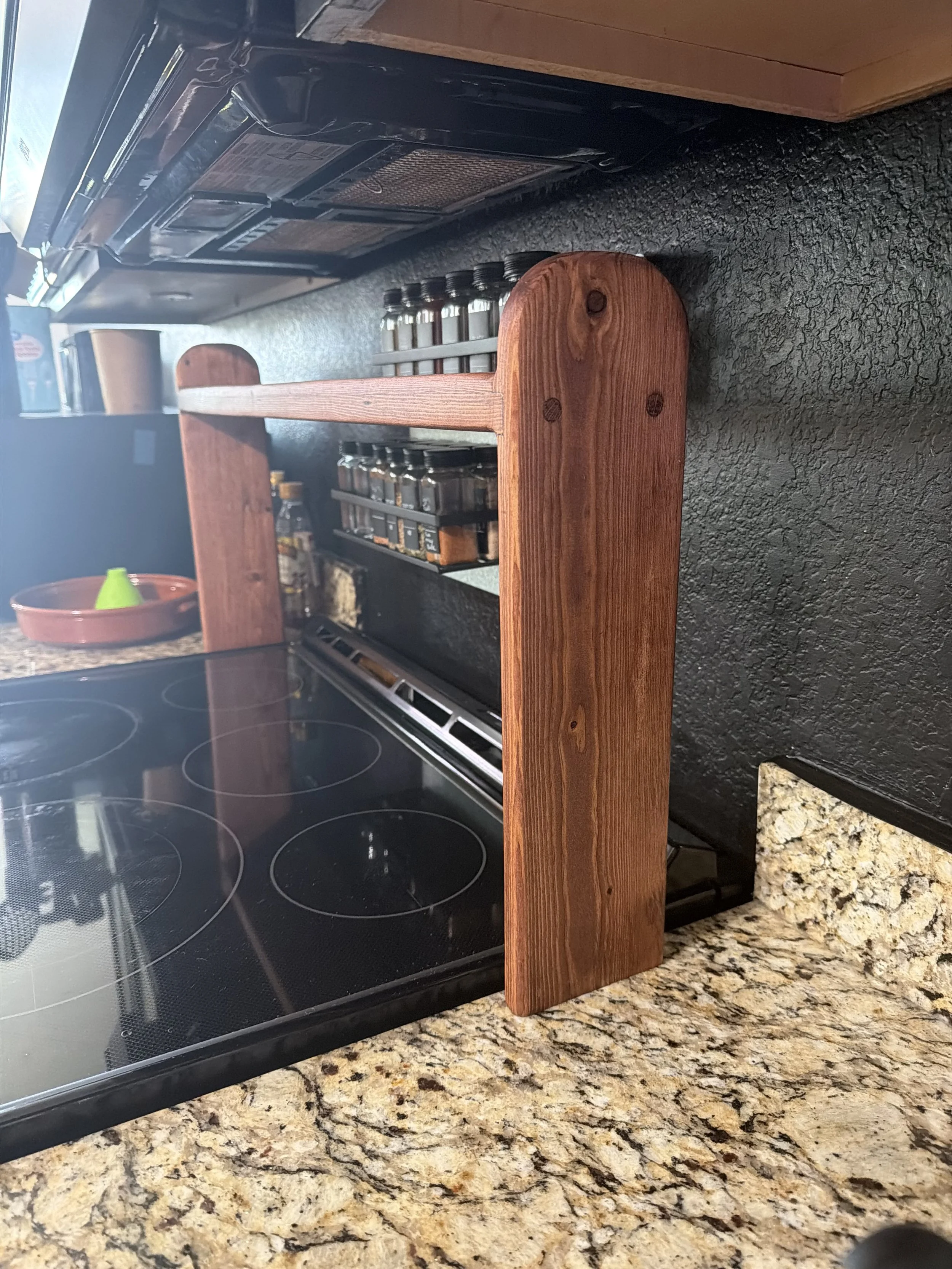 A wooden separator on a granite kitchen countertop in front of a black glass electric stove and a spice rack with jars.