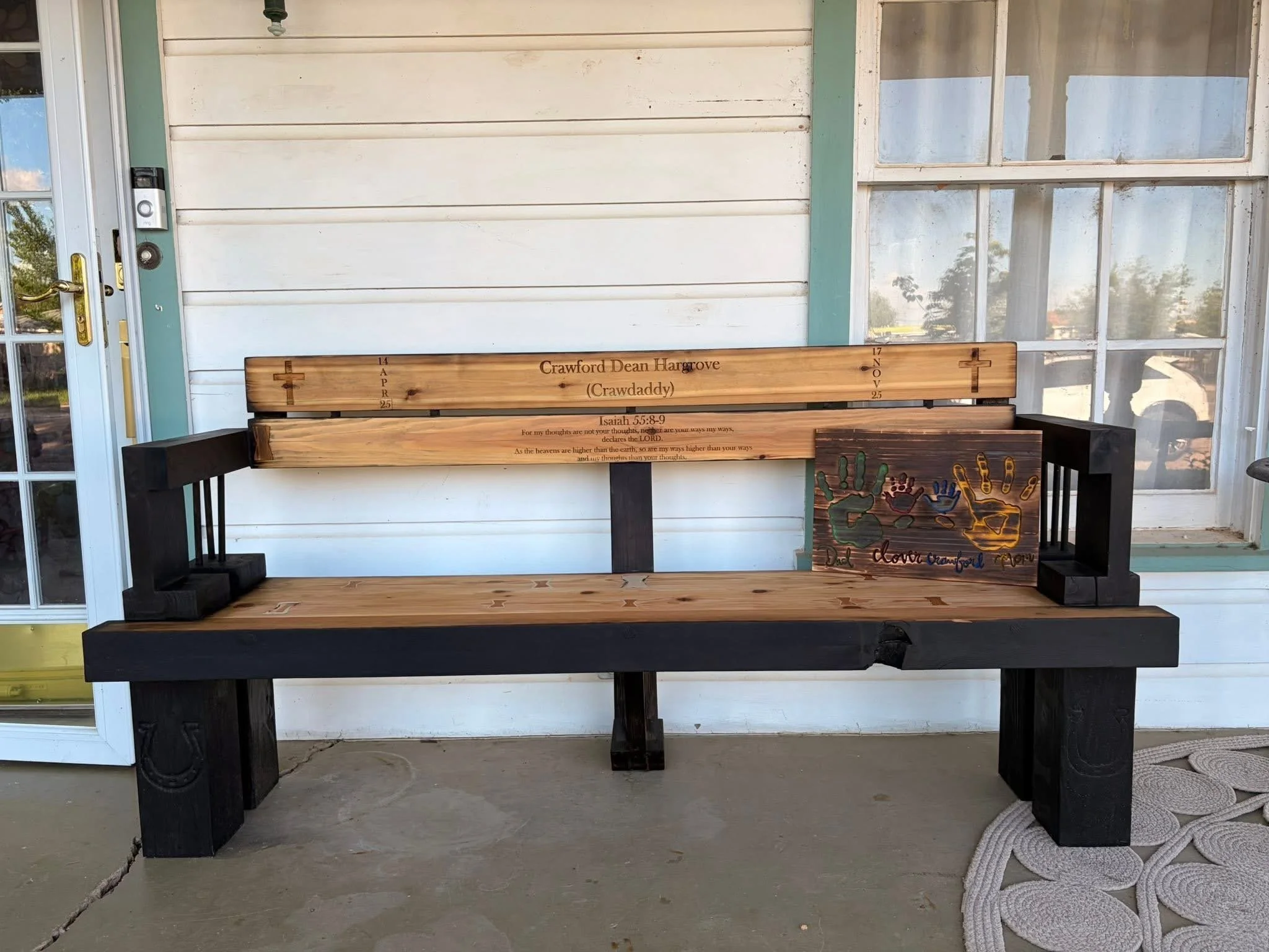 A wooden bench with black armrests and legs sits on a porch in front of a white house with multiple windows. The bench has engraved text commemorating Crawford Dean Hargrove, with dates April 14, 1925, and November 17, 2025, and a Bible verse from Isaiah 55:8-9. To the right of the bench is a colorful piece of artwork with handprints and the phrase "Dad loves Cheyenne". The porch has a light gray floor and a white door frame on the left.