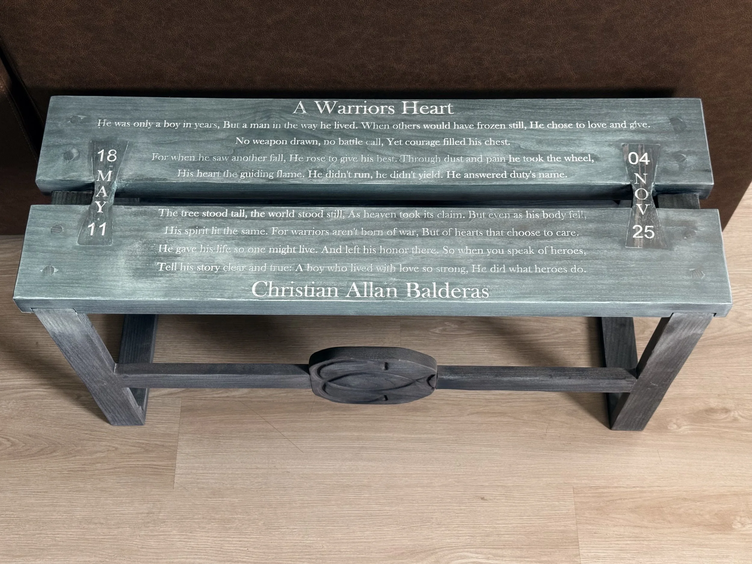 A wooden sign with engraved text and decorative elements, featuring a quote about a warrior's heart and a name at the bottom, placed on a wooden surface.