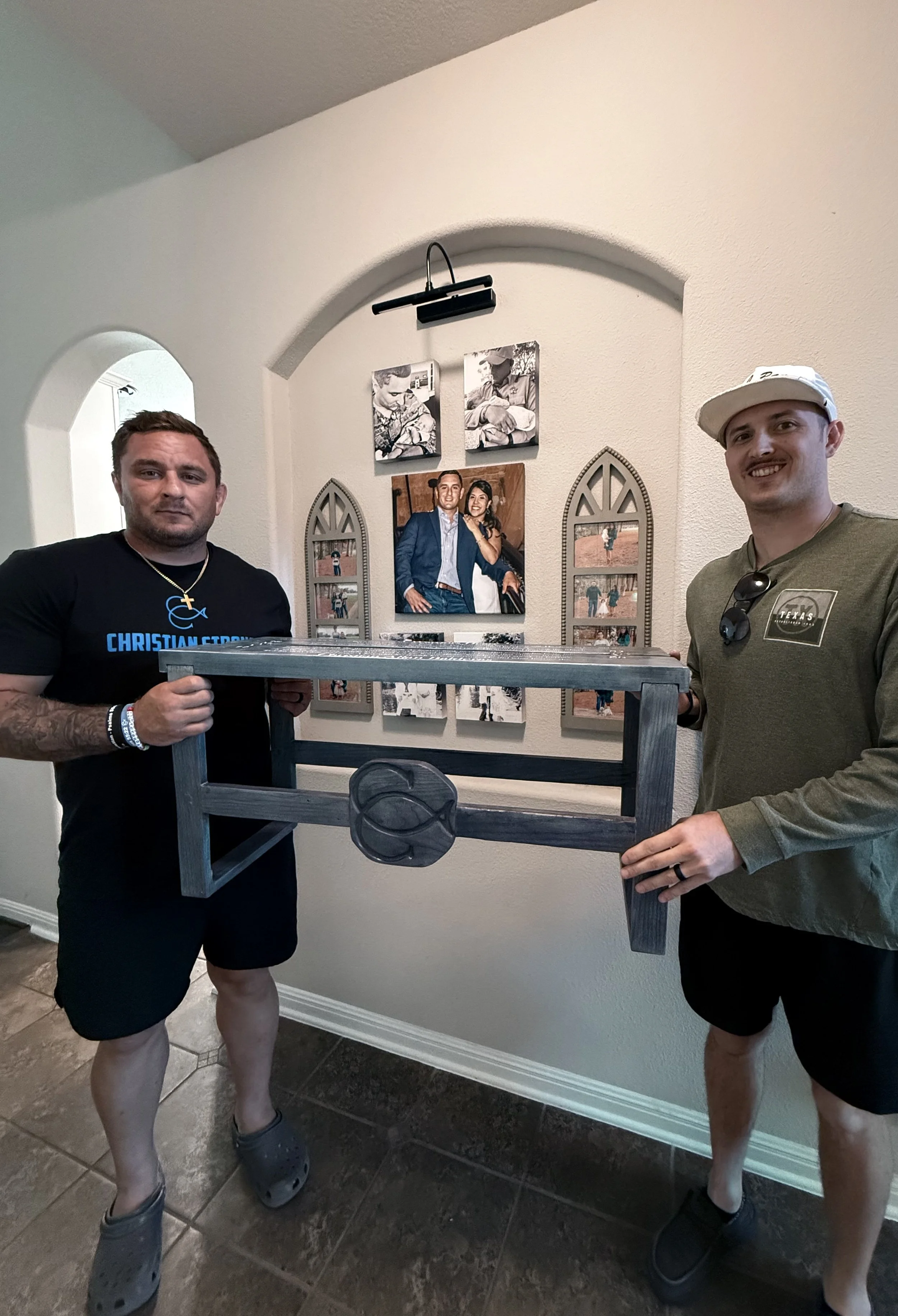 Two men standing indoors holding a large wooden fish-shaped decoration. Both men are dressed casually with shorts and t-shirts, and smiling. The background features framed family photos on the wall.