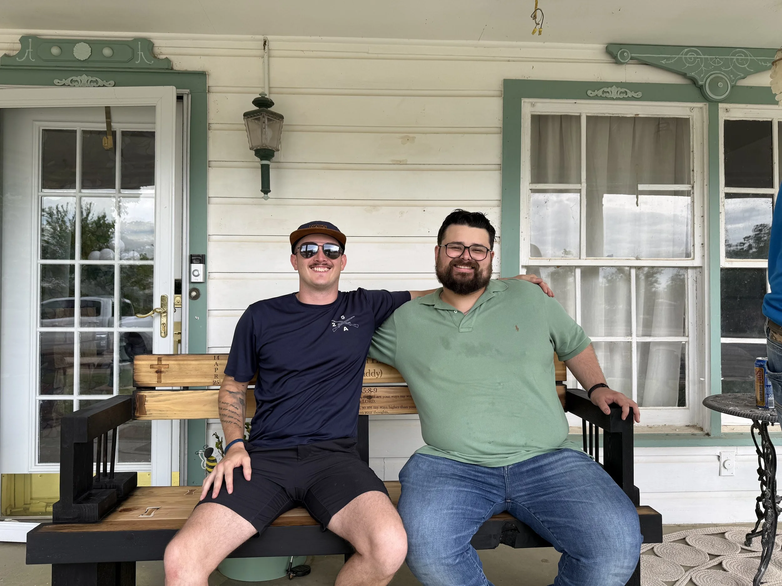 Two men sitting on a front porch, smiling at the camera. One man is wearing sunglasses, a navy blue shirt, and a hat, with tattoos visible on his arm. The other man is wearing glasses and a green polo shirt. Behind them is a white wooden wall with large windows and green trim, with a lamp hanging on the wall and a wooden bench in front of them.