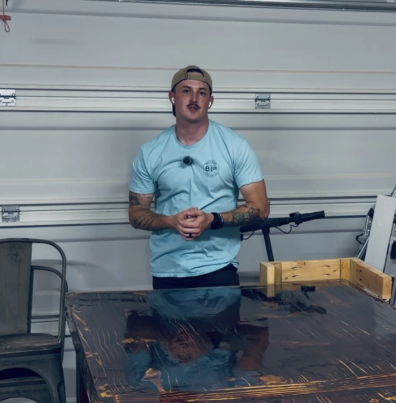 A man with tattoos on his arms, wearing a backwards baseball cap, light blue T-shirt, and wireless earbuds, is leaning against a garage door. He is standing behind a dark, reflective wooden table with a chair to his left and some tools and objects nearby.