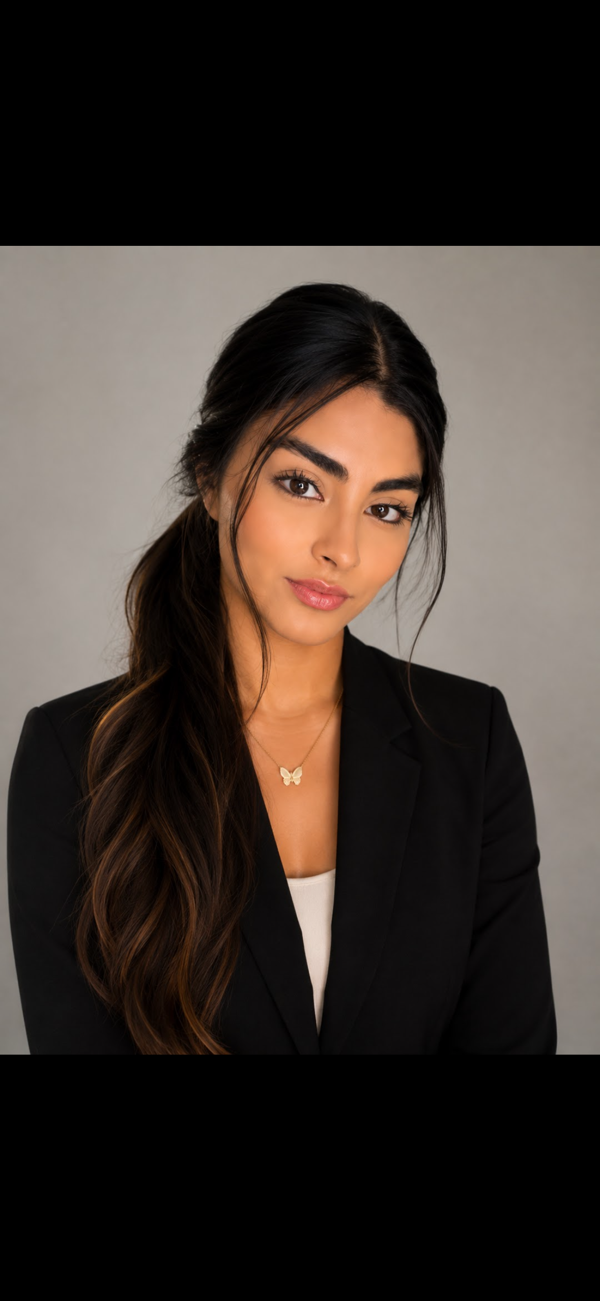 Portrait of a young woman with long dark hair, wearing a black blazer, white top, and butterfly necklace, against a neutral background.