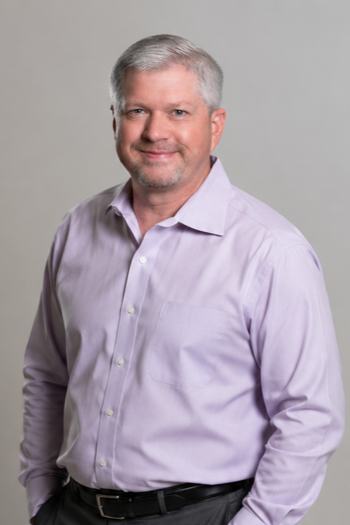 A middle-aged man with short gray hair and a beard, wearing a light purple button-up shirt, smiling, against a plain gray background.