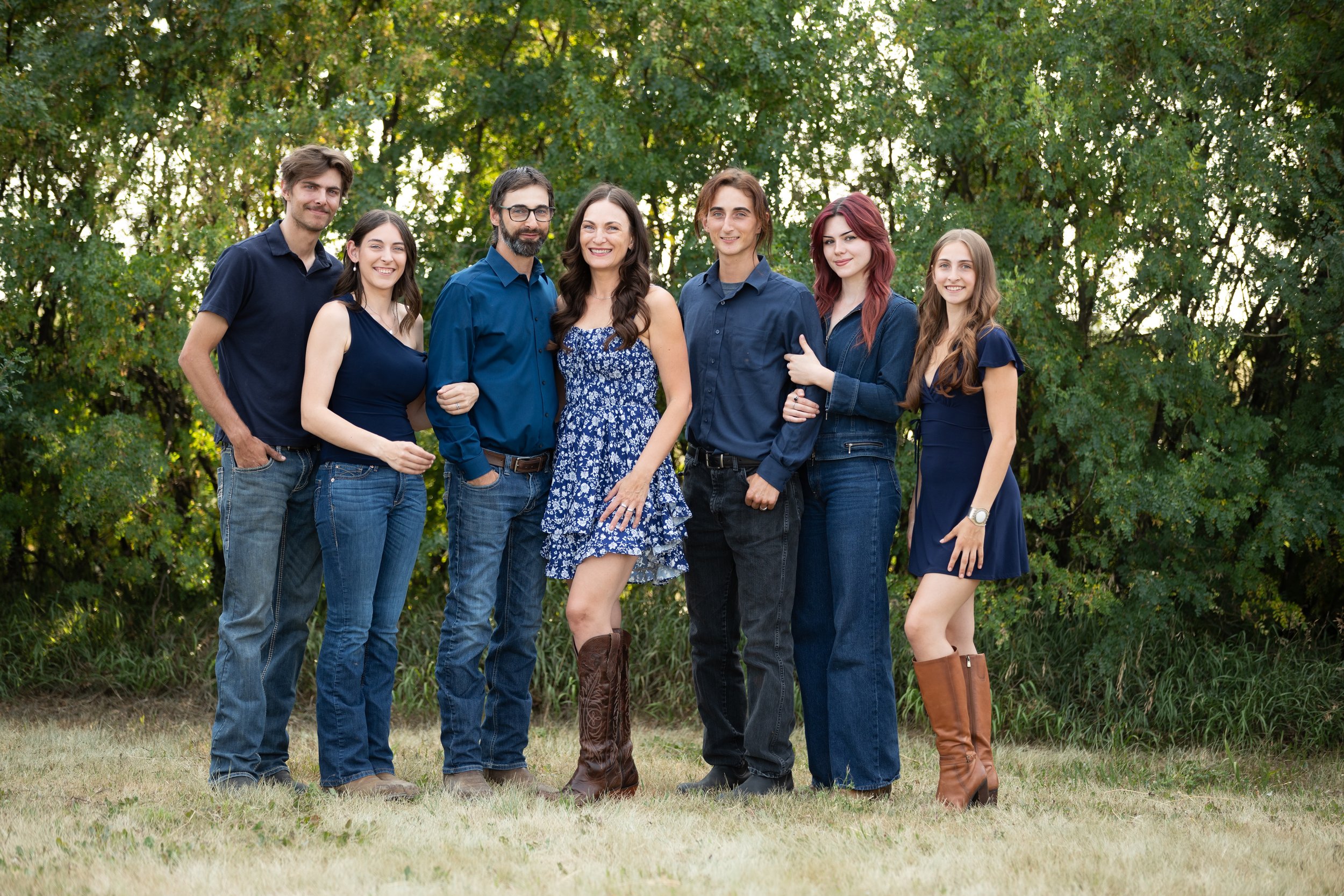Group of seven people standing outdoors in front of green trees, smiling and posing for a photo.