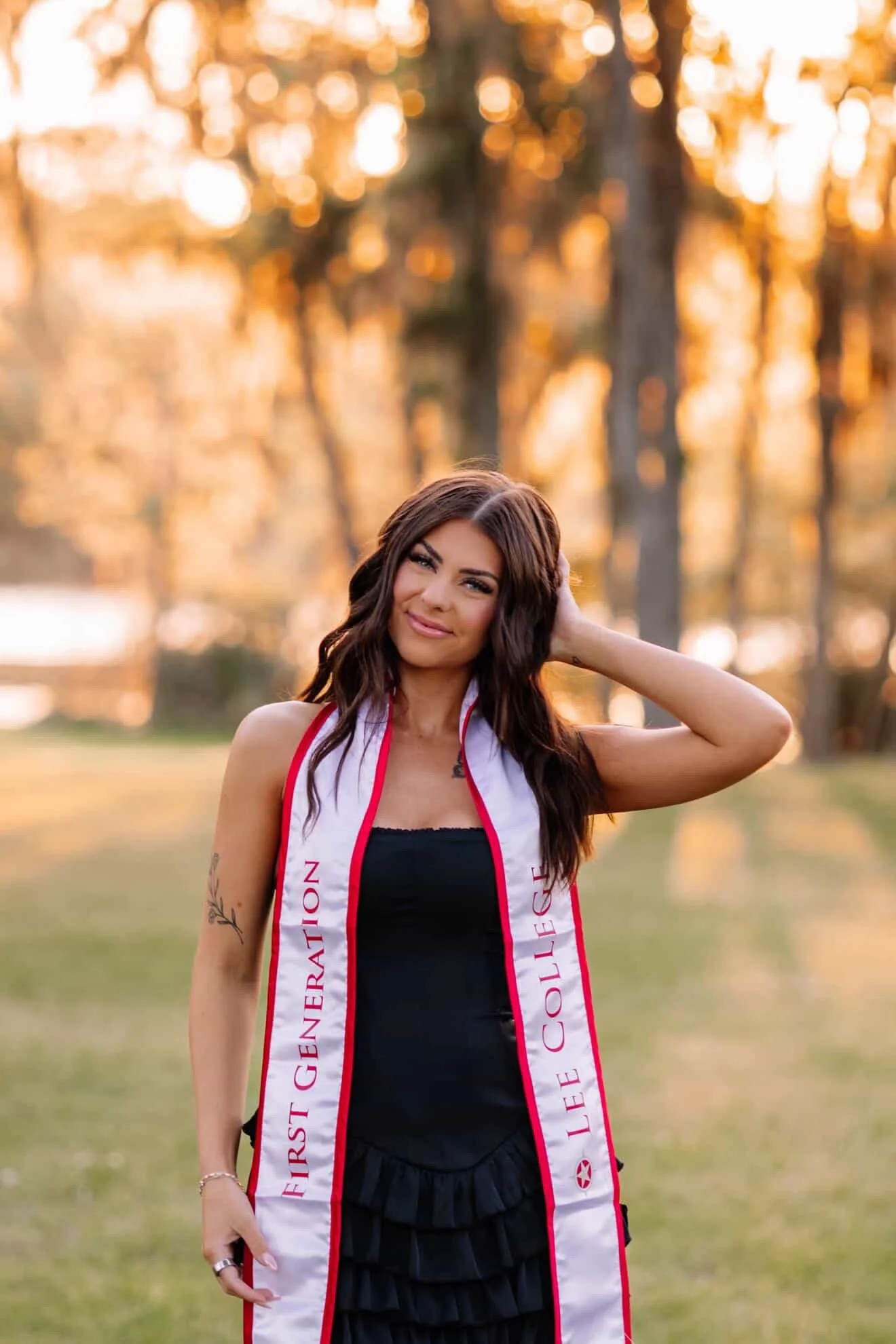 A young woman with long brown hair and tattoos on her arm, smiling and standing outdoors during sunset, wearing a black outfit and a white and red sash with text, in a park with trees in the background.